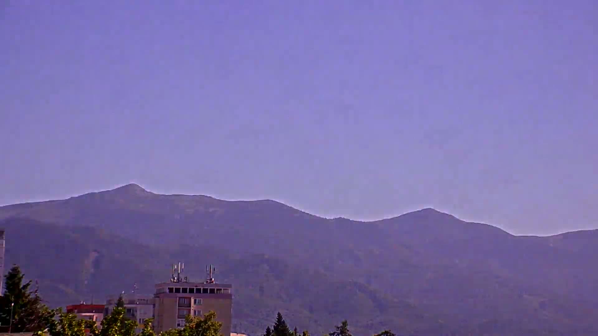 Under a clear, pale purple sky, a range of hazy purple mountains rises behind a collection of low buildings and trees.