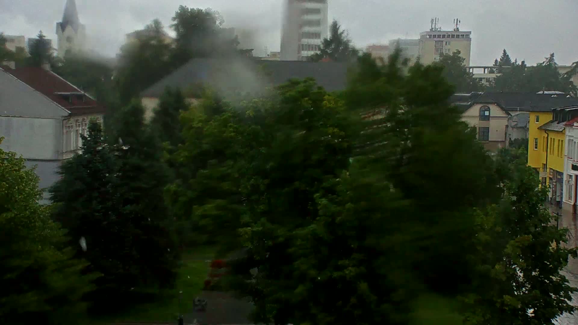 A rain-swept cityscape is seen from an elevated vantage point, showing buildings, trees violently swaying in the wind, and a church spire partially obscured by the downpour.