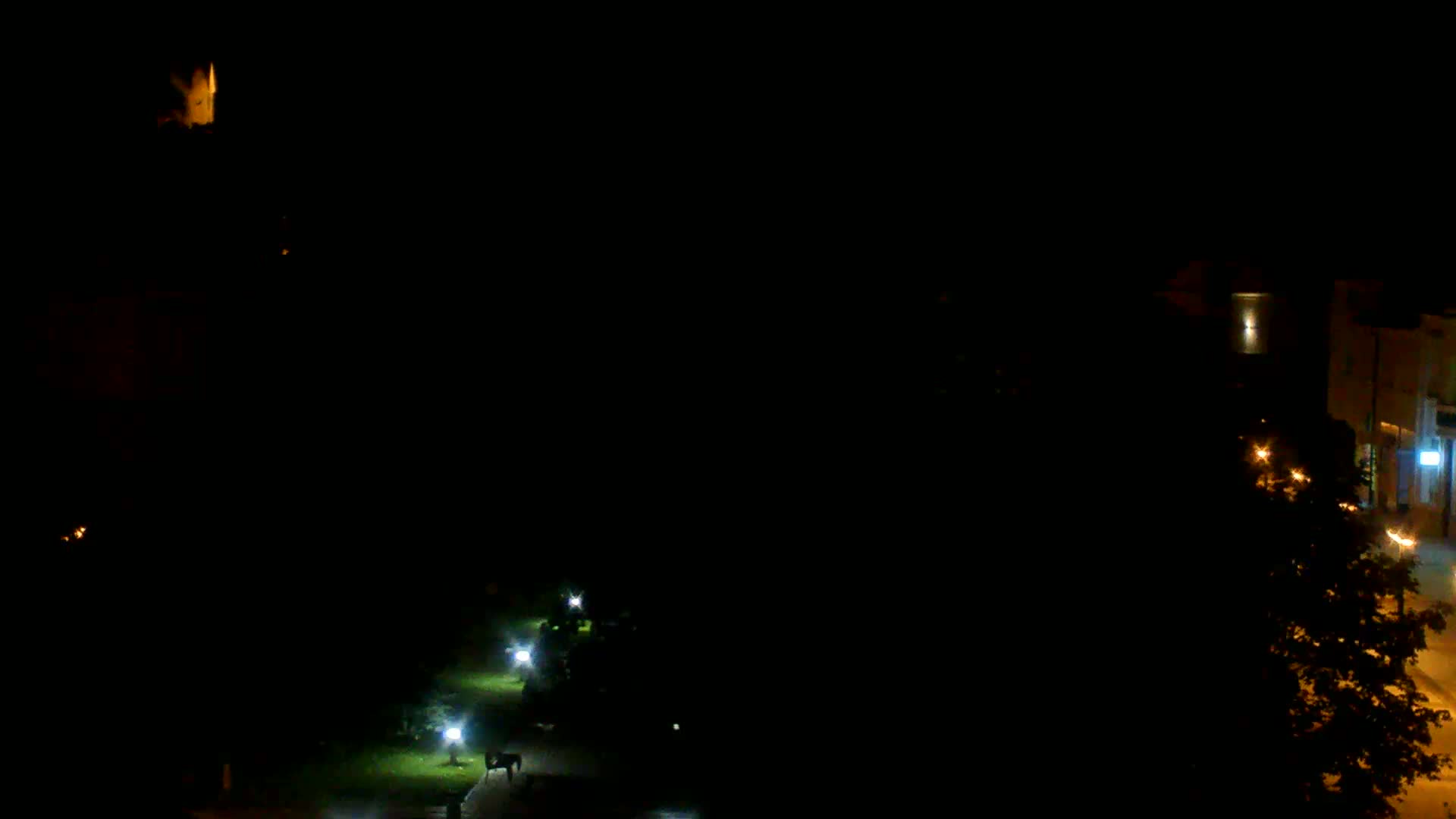 A nighttime, overhead view shows a dark park with a few illuminated paths and a building with lights visible in the distance.