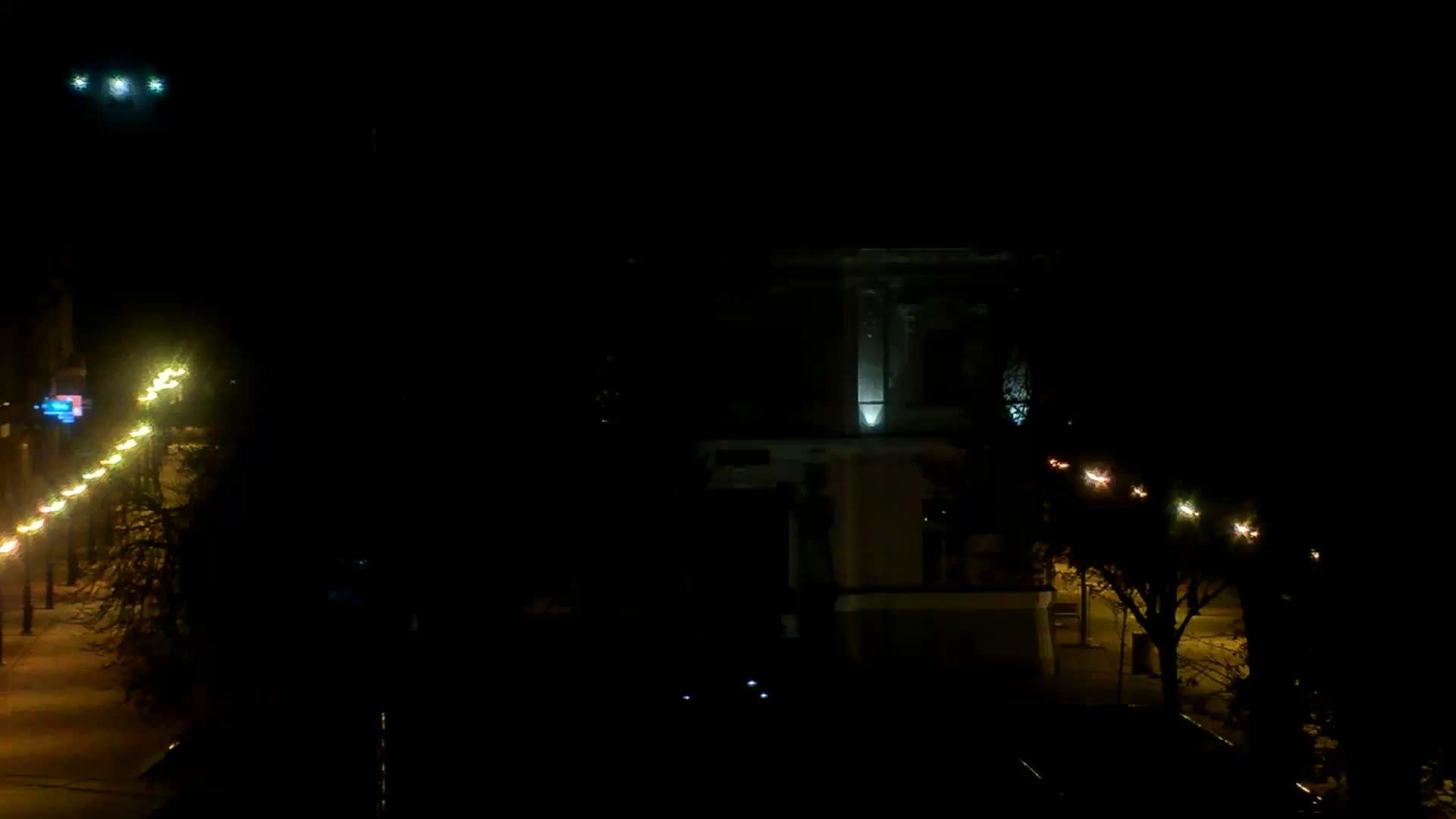 A dark outdoor scene at night shows rows of streetlights illuminating pathways and a brightly lit building in the distance under clear skies.