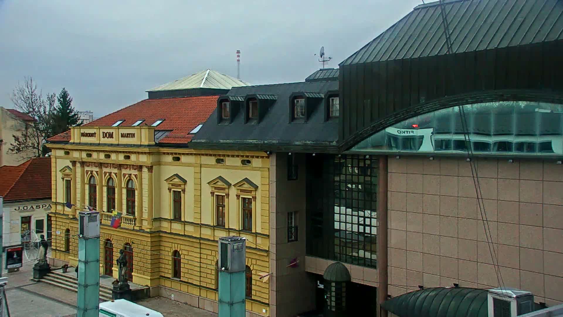 An urban street scene is depicted under an overcast sky, showcasing an ornate yellow building with a red tiled roof juxtaposed with modern structures featuring dark metal roofs and glass facades.
