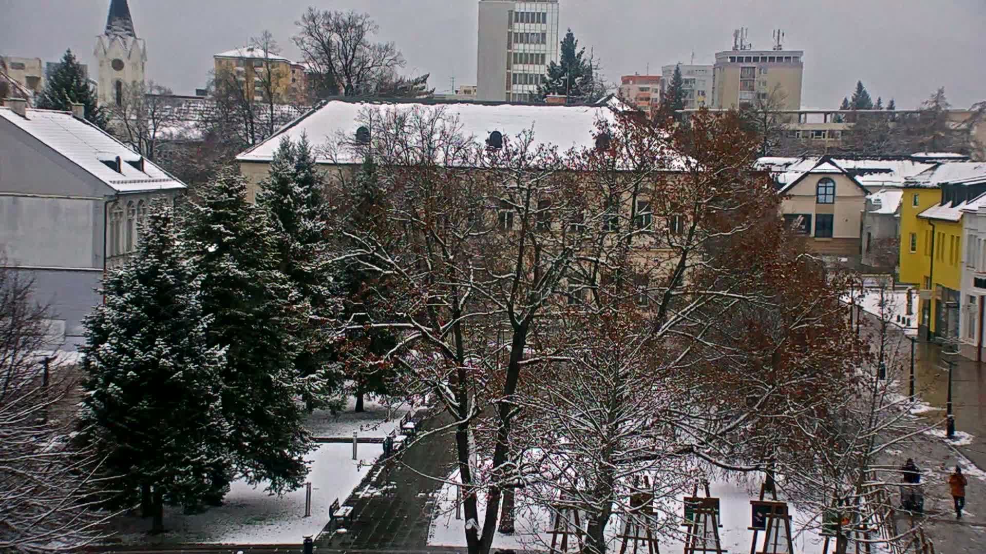 A snow-covered townscape unfolds under a grey, overcast sky, revealing buildings with white roofs, a mix of bare and evergreen trees dusted with snow, and a few individuals traversing a partially cleared path.