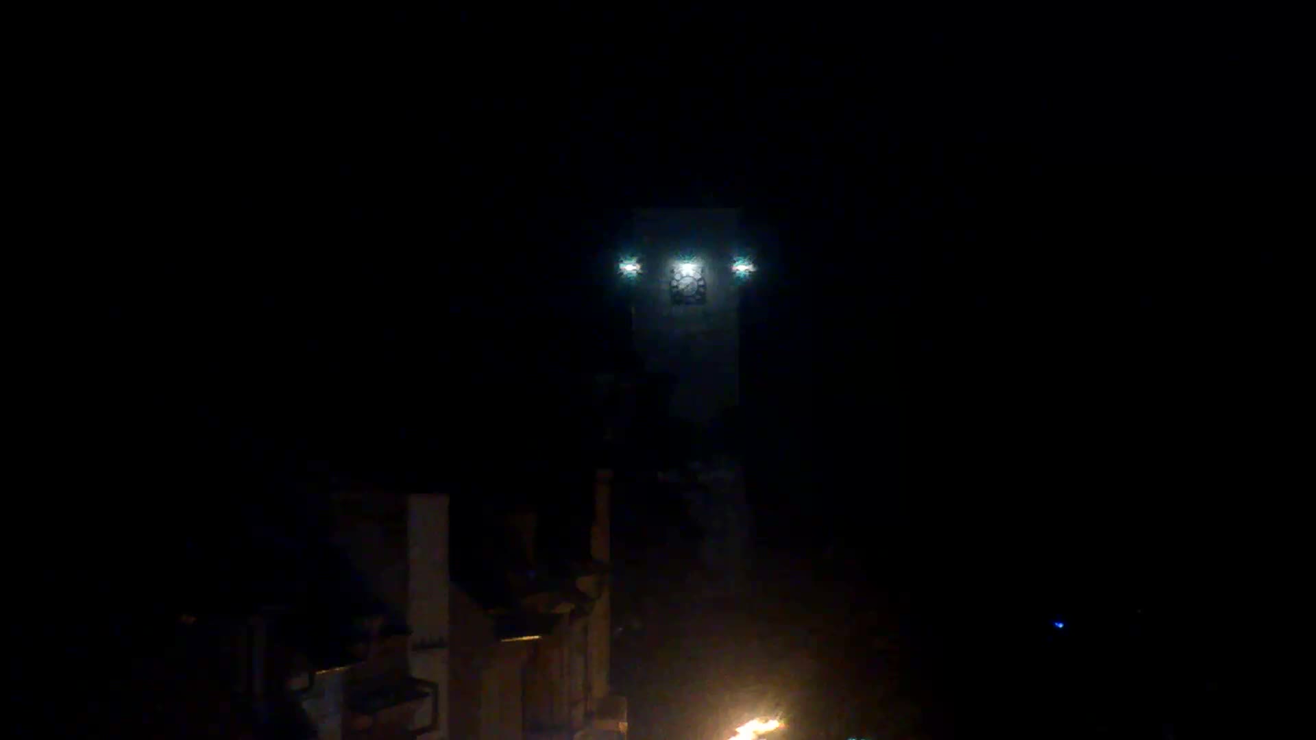 A tall, dimly lit tower with three bright lights and a visible clock face stands in the distance on a dark night, with a vibrant fire glowing in the lower right foreground.
