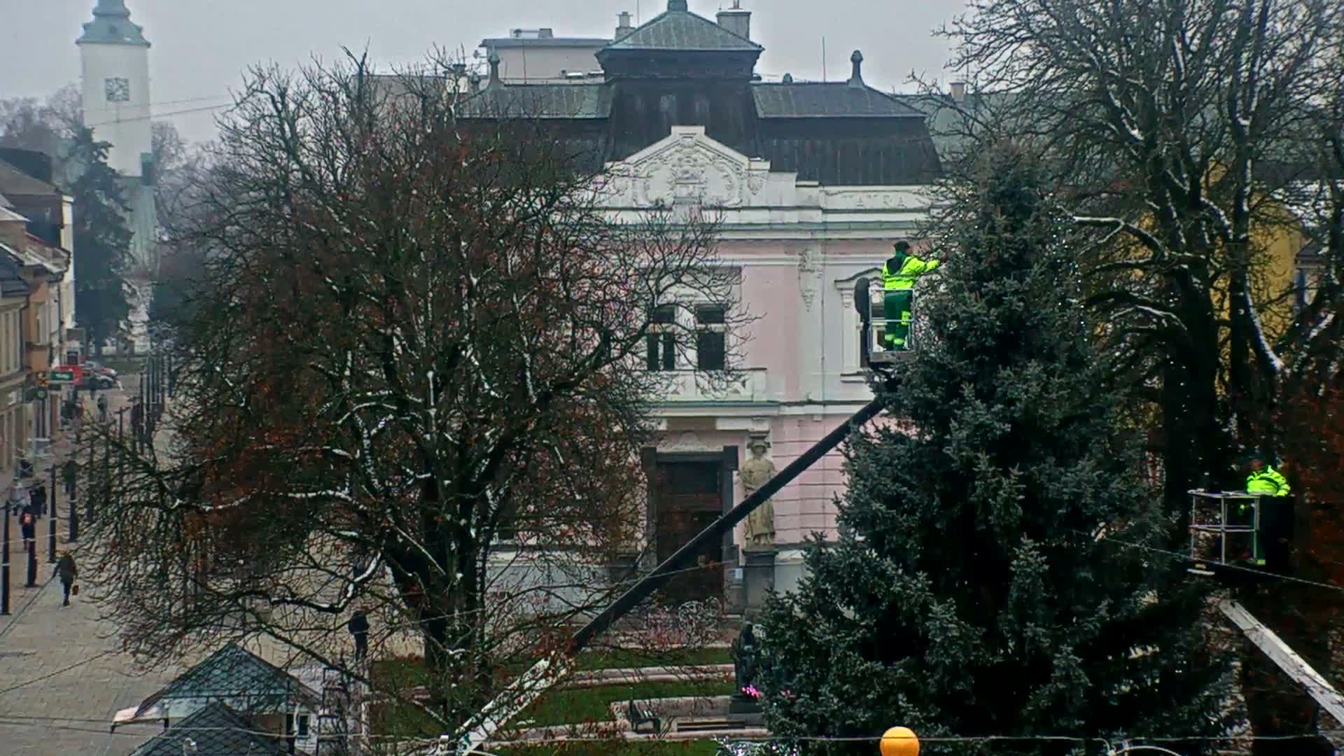 On an overcast, wintry day with remnants of snow on the bare trees, two workers in neon green jackets are decorating a large evergreen tree with lights from a lift, set against an ornate, light pink building and a bustling street with a church tower visible in the distance.