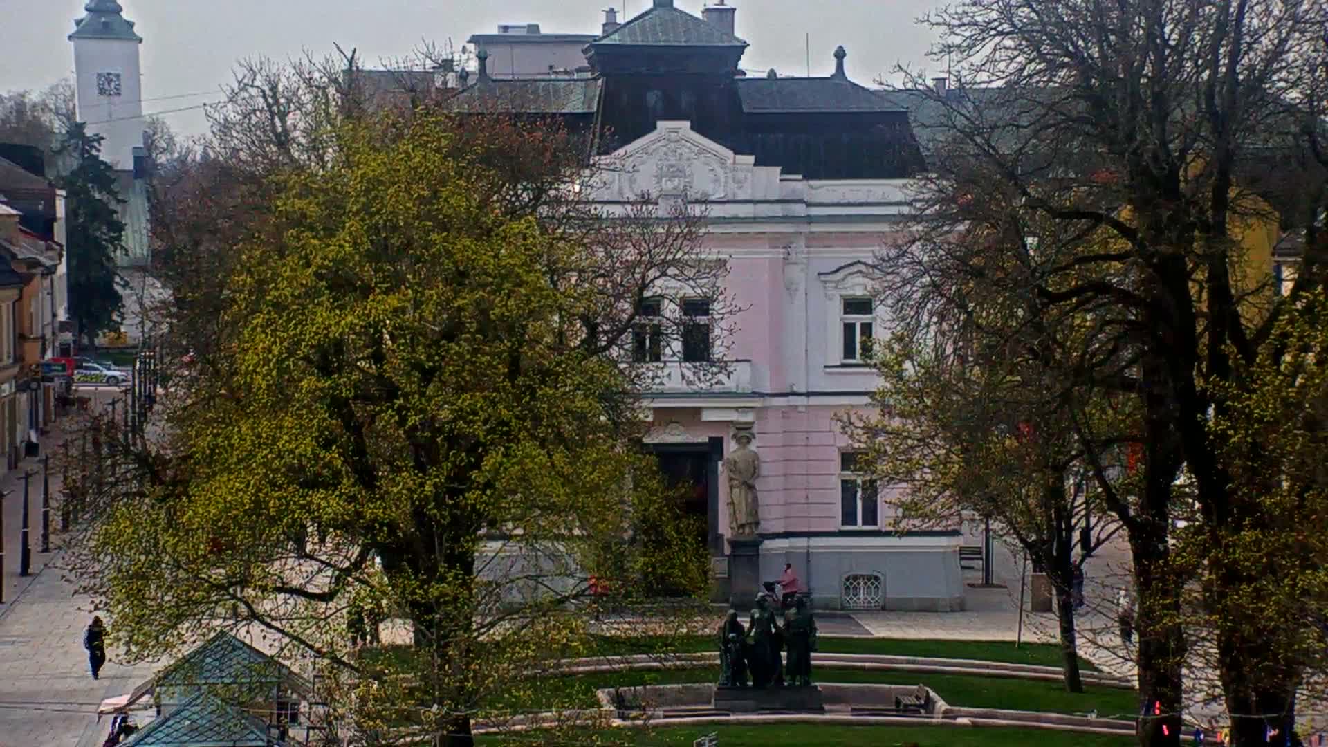 On an overcast, wintry day with remnants of snow on the bare trees, two workers in neon green jackets are decorating a large evergreen tree with lights from a lift, set against an ornate, light pink building and a bustling street with a church tower visible in the distance.