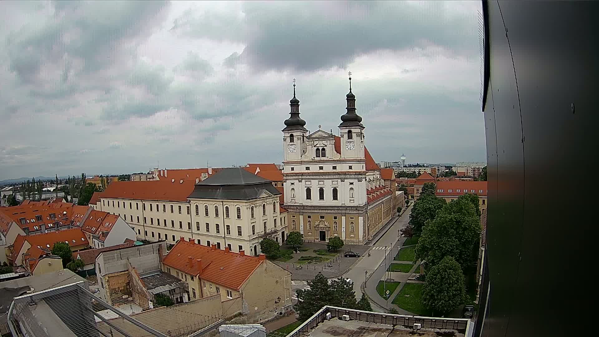 A large church with two towers is situated in a city square, surrounded by other buildings under a cloudy sky.
