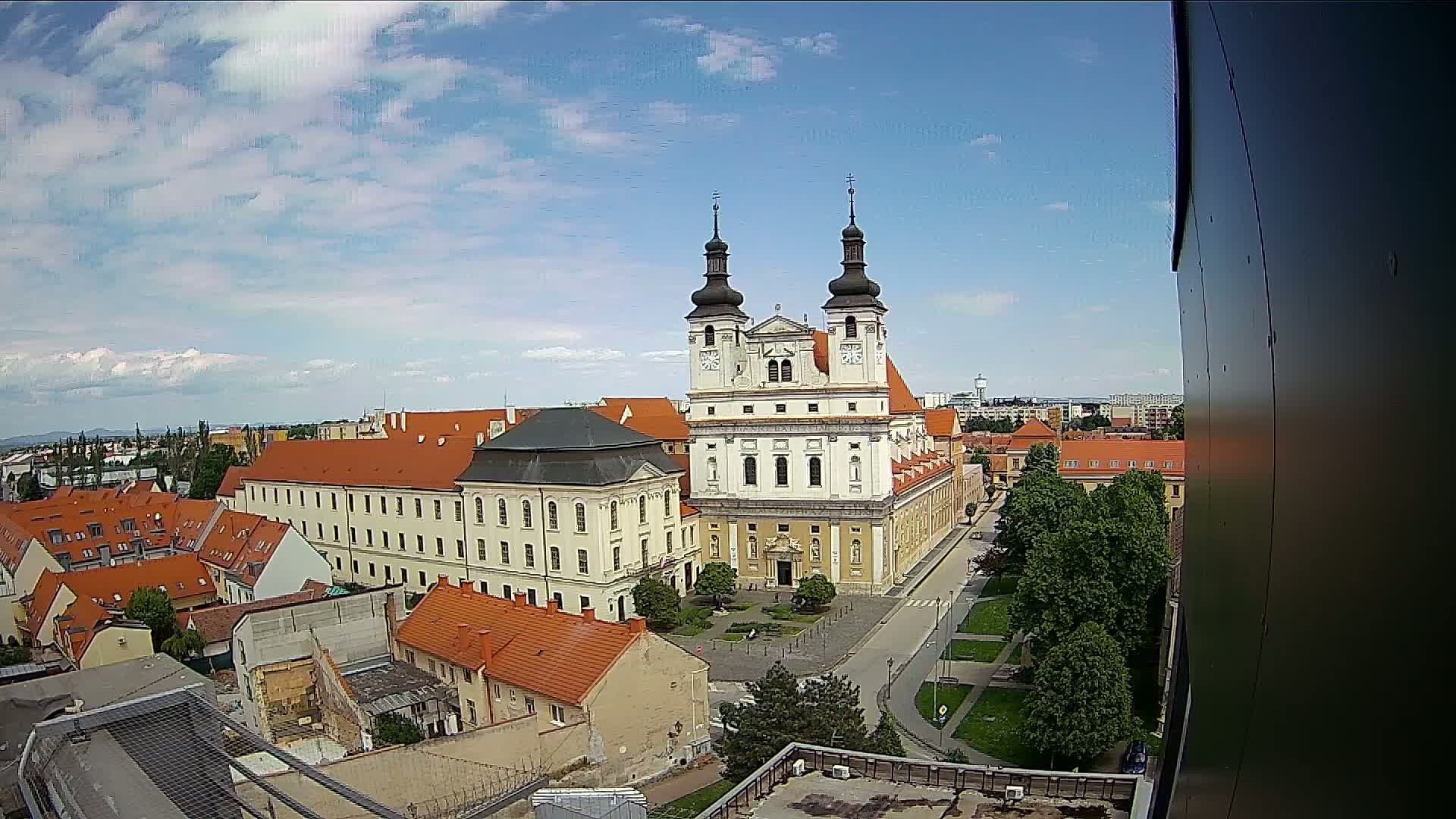 A large white church with two towers is surrounded by red-roofed buildings and green trees under a partly cloudy sky.