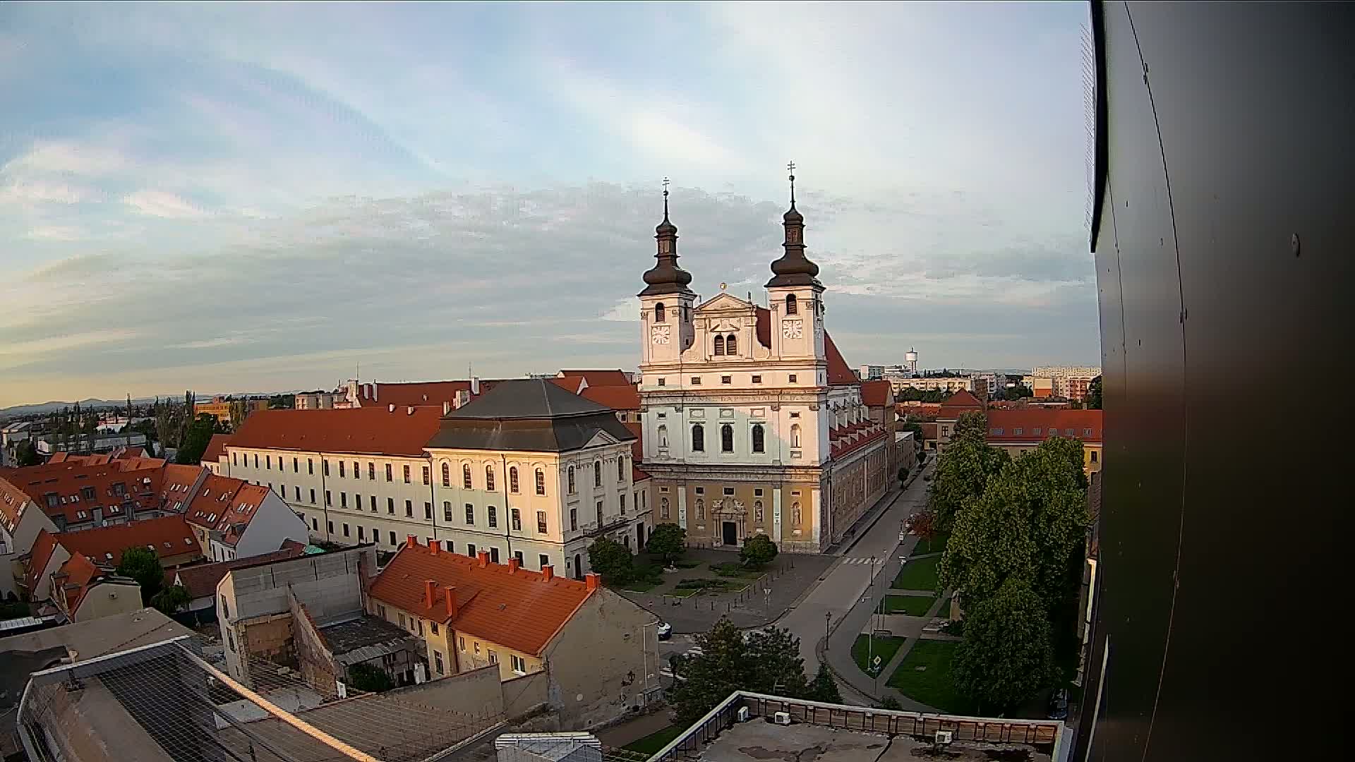 An elevated view of a city on a partly cloudy day shows a large church with two towers in the center of a square surrounded by buildings.