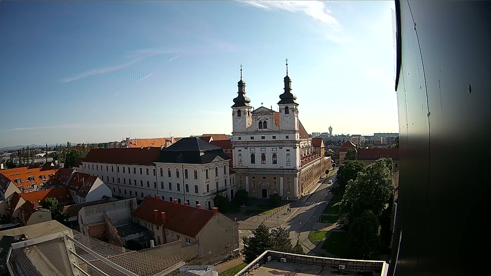 A high-angle, long shot on a clear, sunny day shows a large white church with twin towers, surrounded by other buildings, and a street with trees.