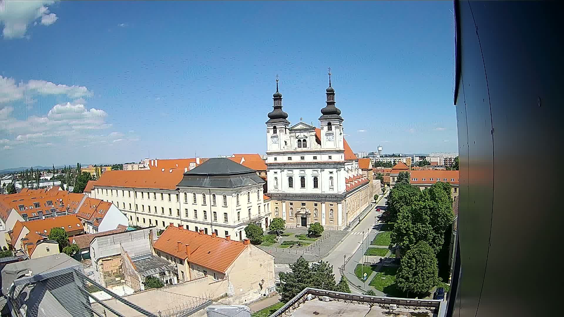 An elevated view of a city on a sunny day shows a large church complex surrounded by buildings with red-tiled roofs and green trees.