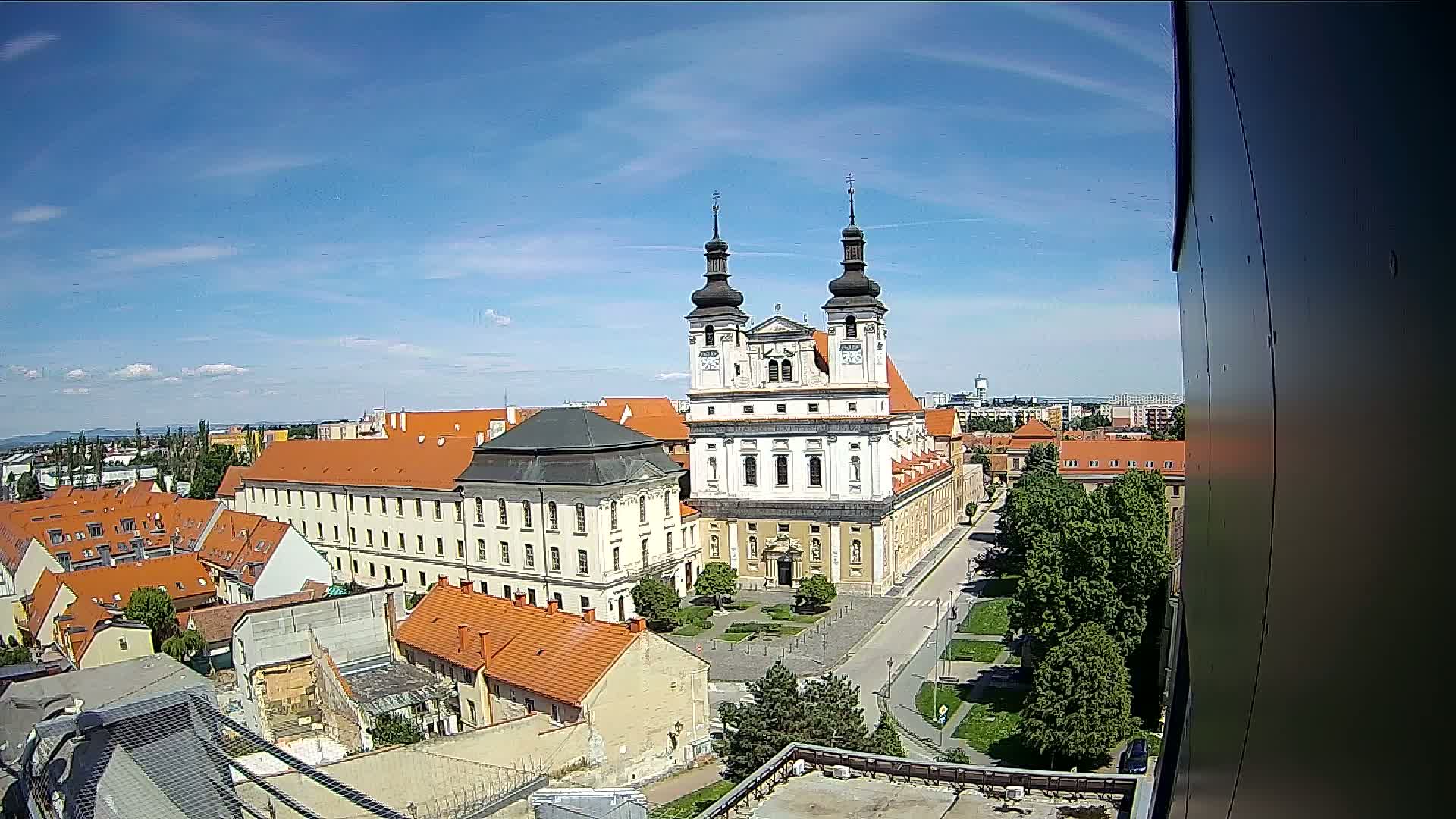 A large white church with twin towers stands in a city square on a sunny day, surrounded by red-roofed buildings and green trees.
