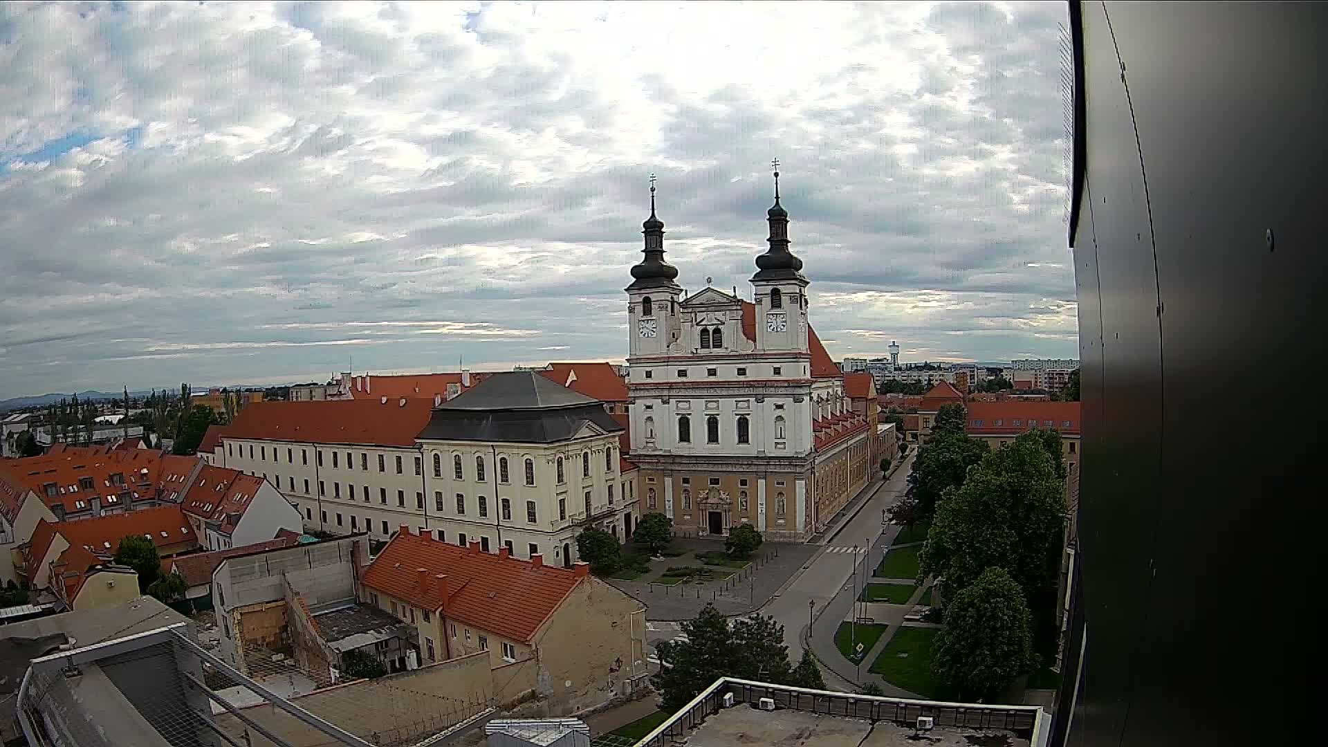 A high-angle view of a city on a partly cloudy day shows a large white church with twin towers at its center, surrounded by smaller buildings with red tile roofs.