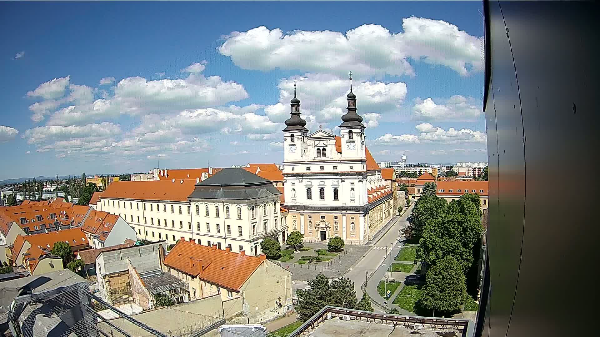 A large, light-colored church with two towers stands prominently amidst a cluster of red-roofed buildings under a partly cloudy blue sky.