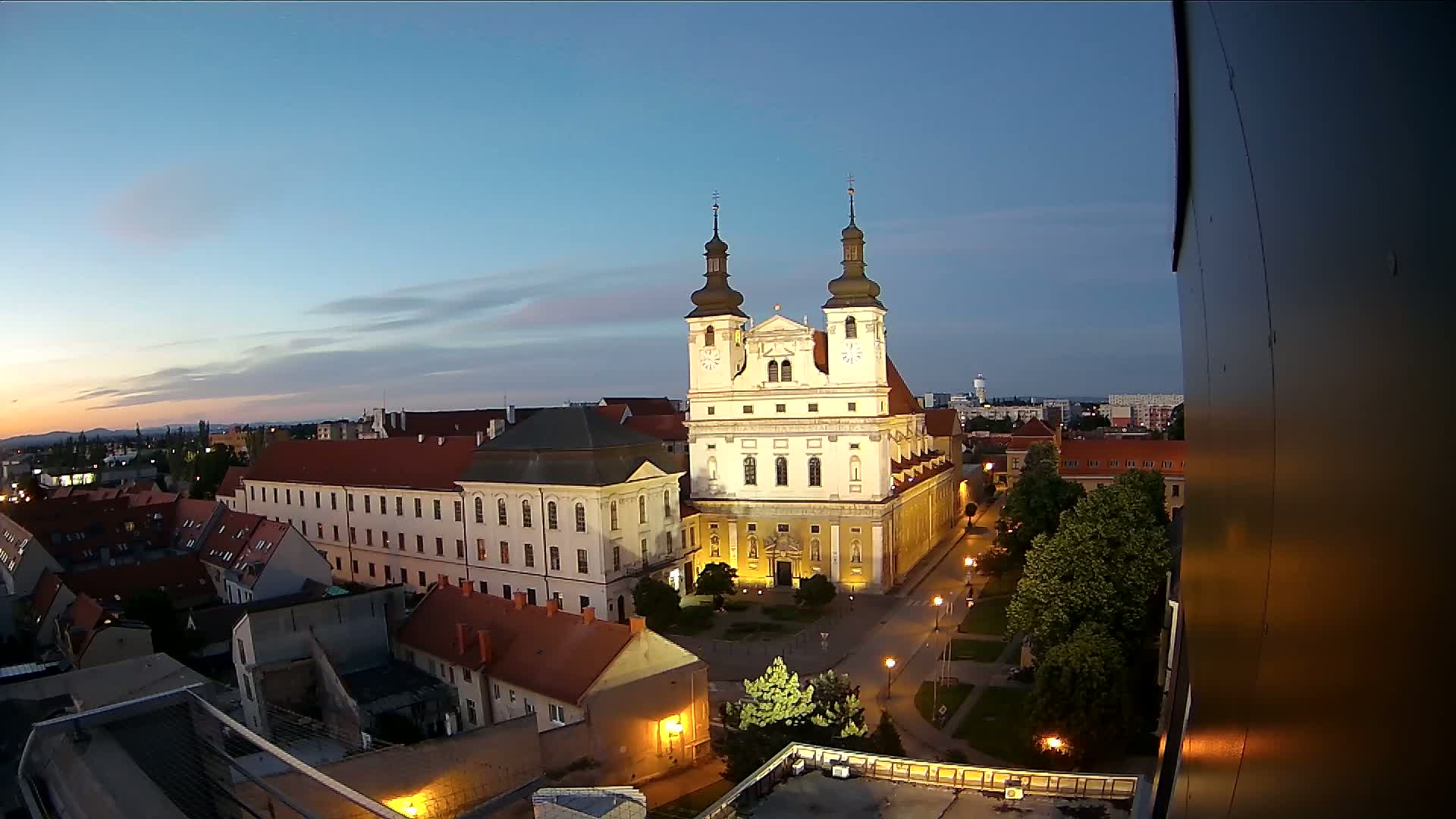 A large, illuminated church stands in a city at twilight, surrounded by smaller buildings and trees under a clear, dusky sky.