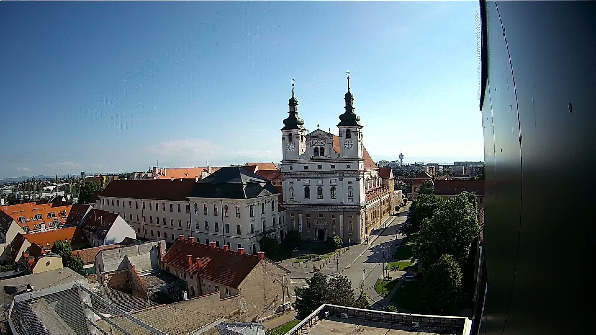 A large, light-colored cathedral with two towers stands in the center of a town square surrounded by smaller buildings under a clear, sunny sky.