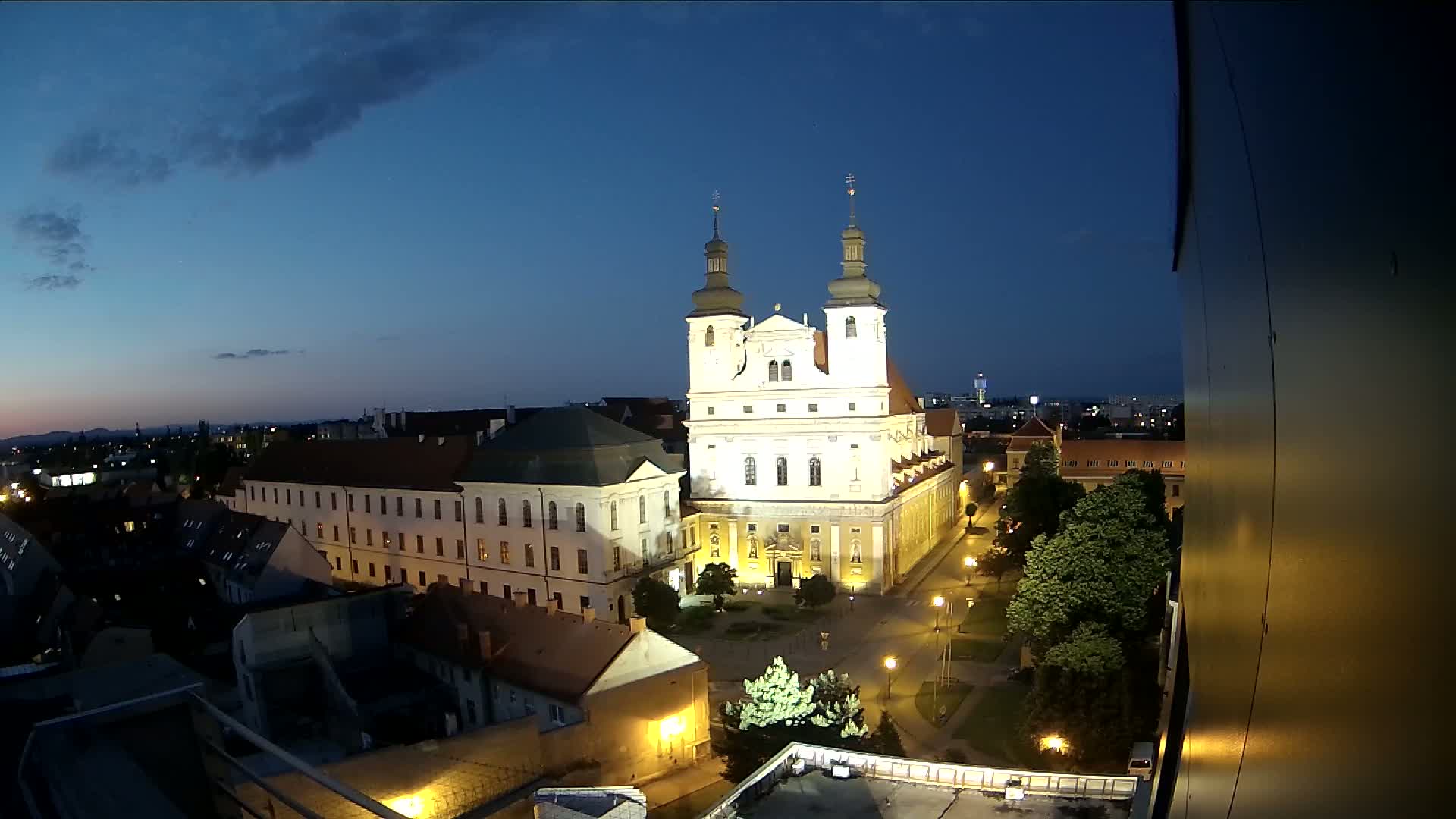 A large, illuminated church stands in the center of a city at twilight, surrounded by smaller buildings and a park, under a mostly clear sky.