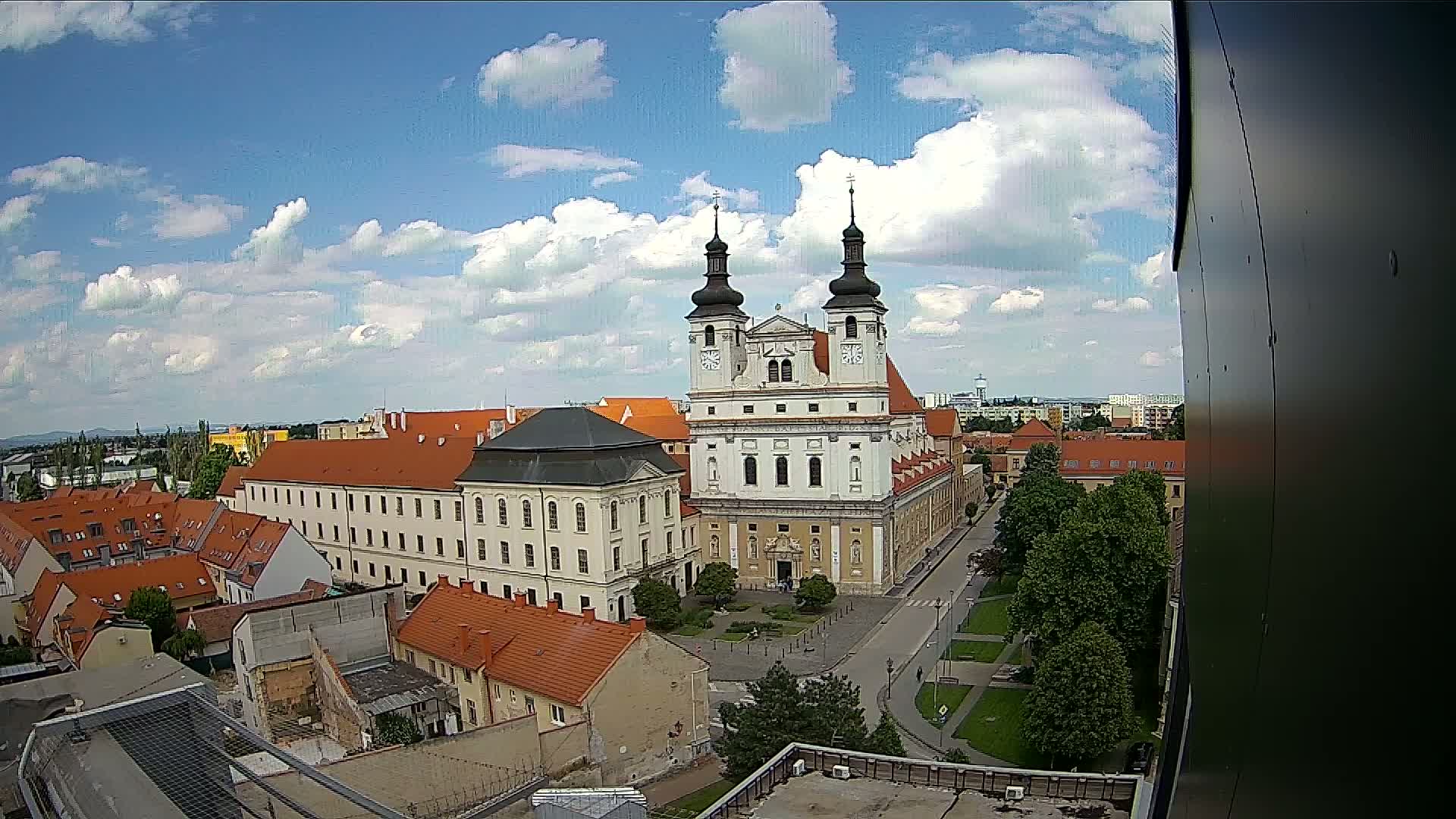 A large, light-colored church with twin towers and red-roofed buildings surrounds a town square under a partly cloudy sky.