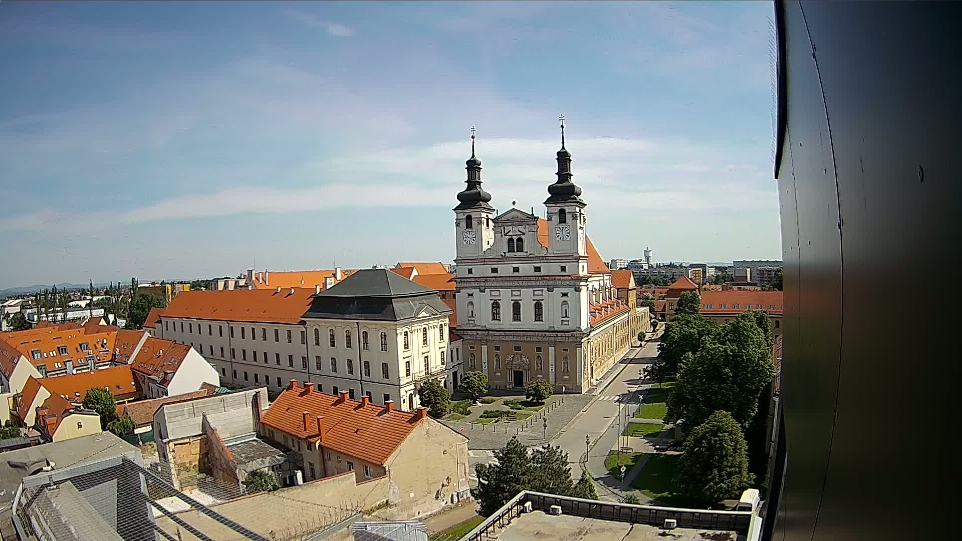 A large church with twin towers stands in the center of a town under a mostly sunny sky, surrounded by low buildings with orange roofs and green trees.