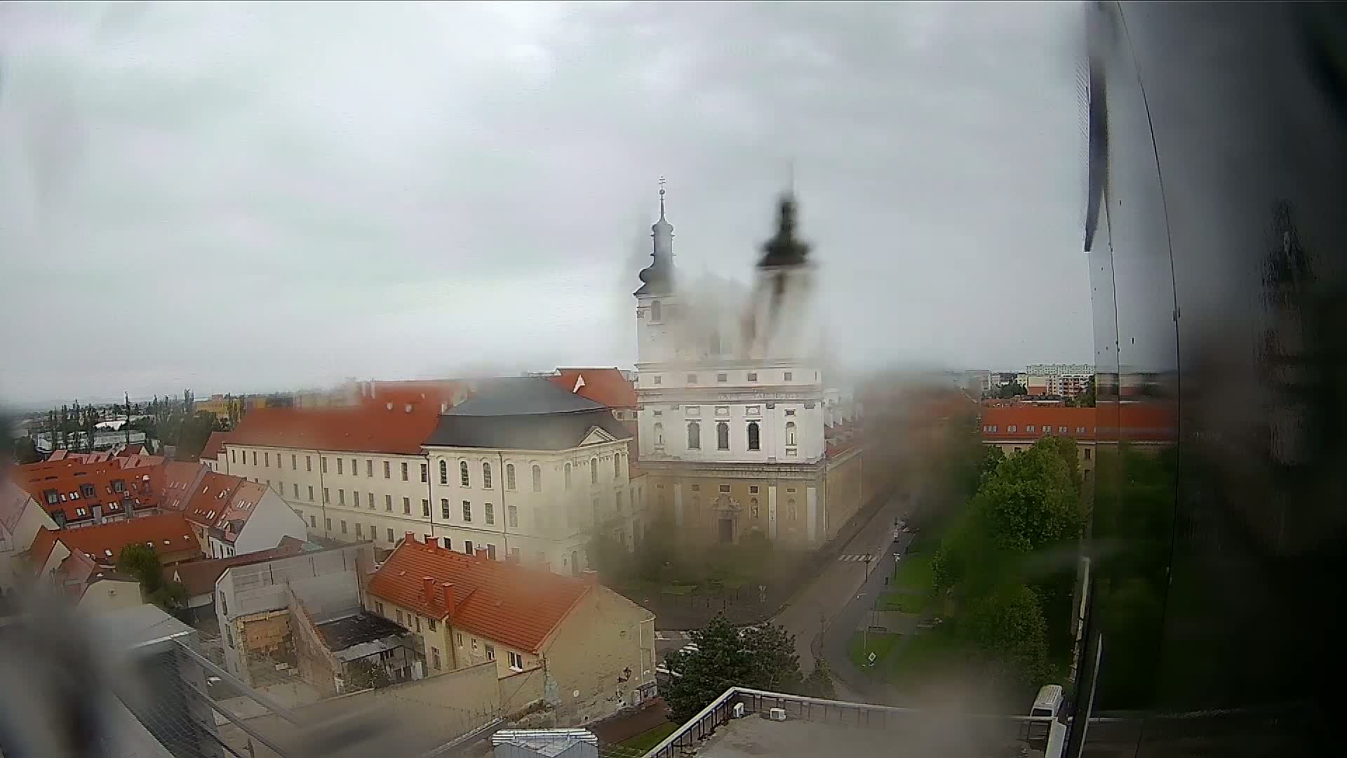 A rain-streaked view from a window shows a European-style city square with a large church and surrounding buildings on an overcast day.