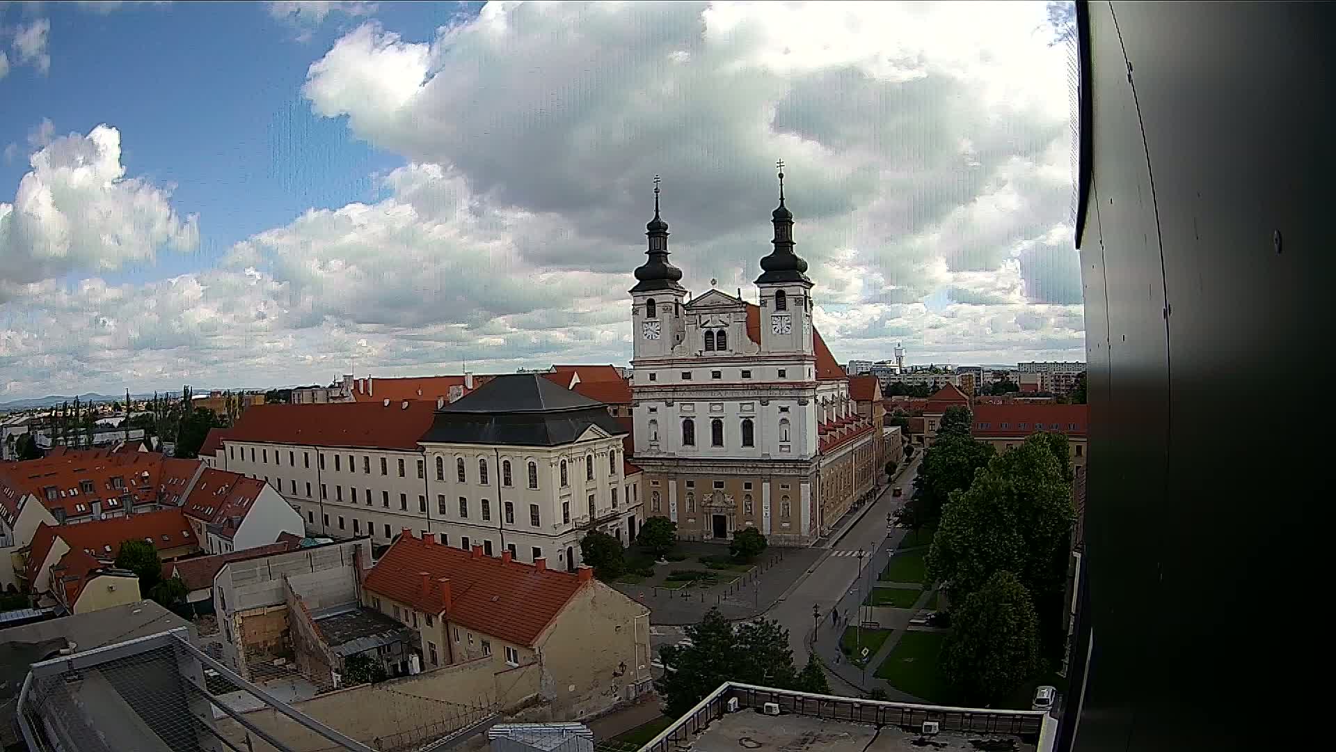 A partly cloudy sky overlooks a European city square centered on a large white church with two dark spires, surrounded by red-roofed buildings and green trees.