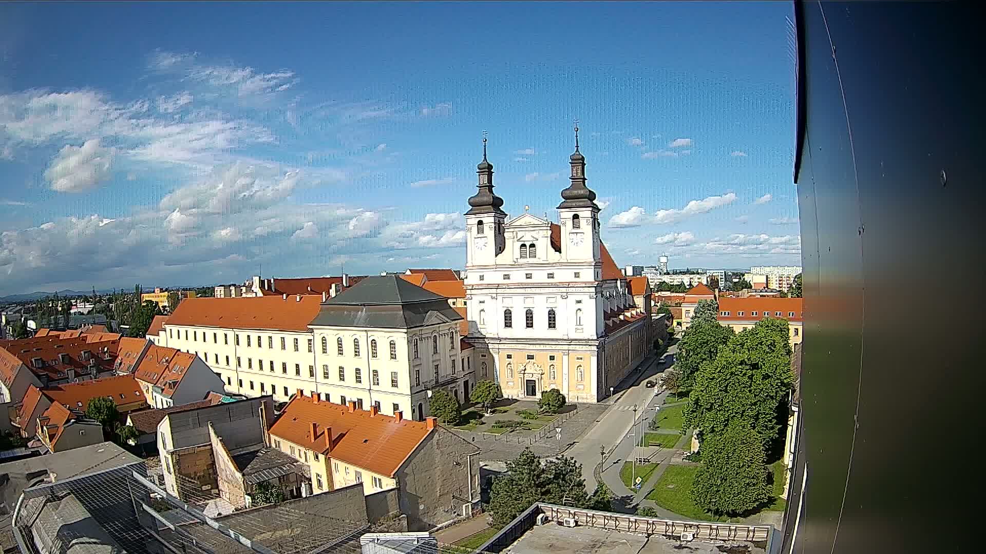A large church with two towers stands in the center of a town under a mostly sunny sky with scattered clouds.