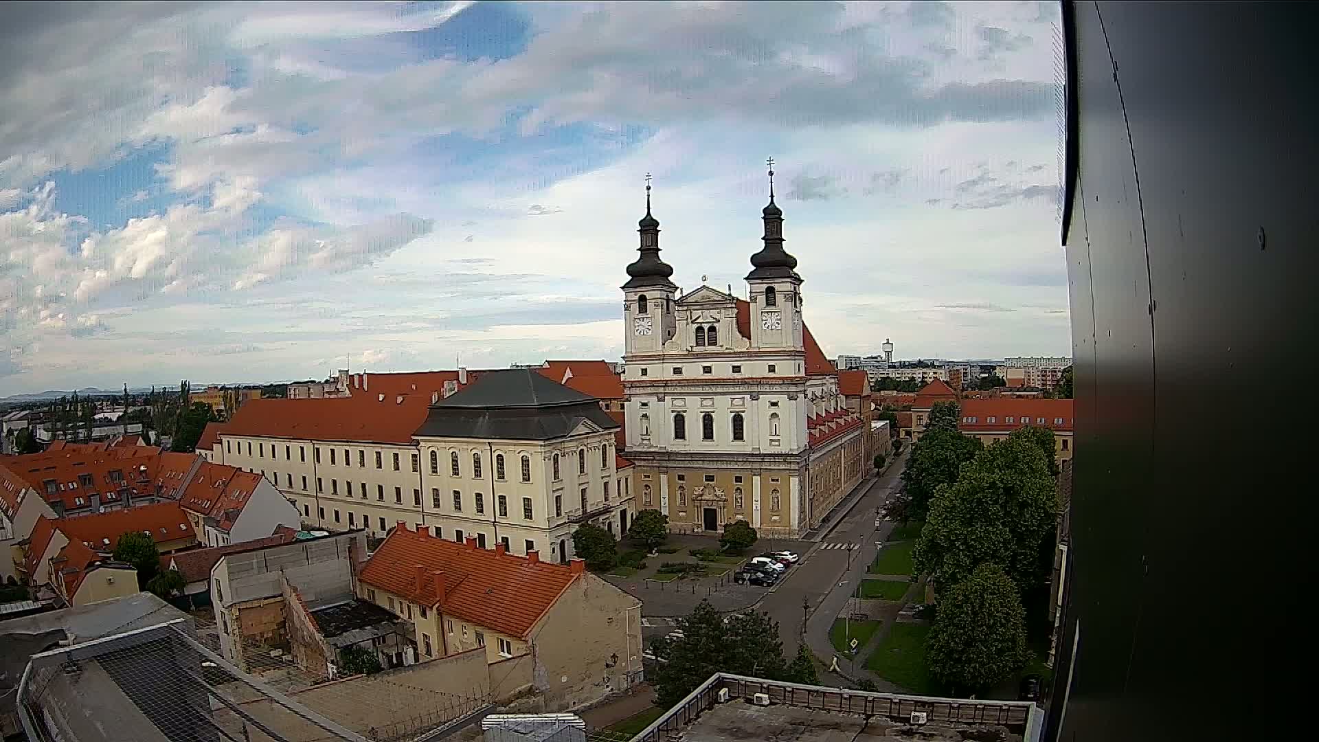 An elevated view of a European city on a partly cloudy day shows a large church and surrounding buildings, with trees and a street visible in the foreground.