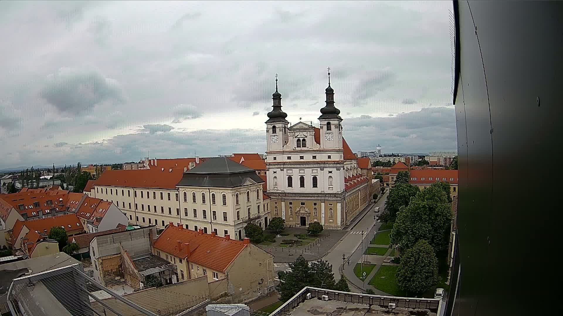 An elevated view of a city under a cloudy sky shows a large church with two towers at its center, surrounded by buildings with red-tiled roofs and green spaces.