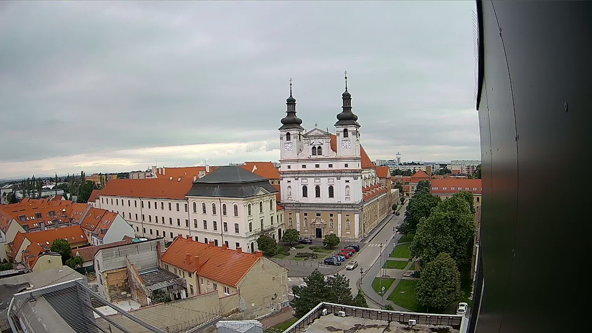 An elevated view of a European city on an overcast day shows a large white church with two towers, surrounded by buildings with red-tiled roofs and green trees.