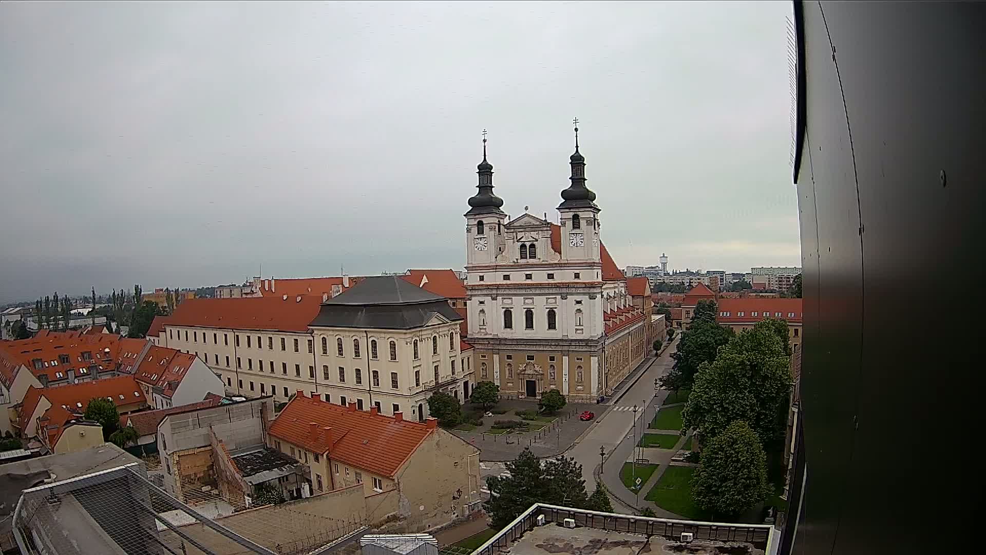 A large church with two towers stands in the center of a town under an overcast sky, surrounded by other buildings and green spaces.