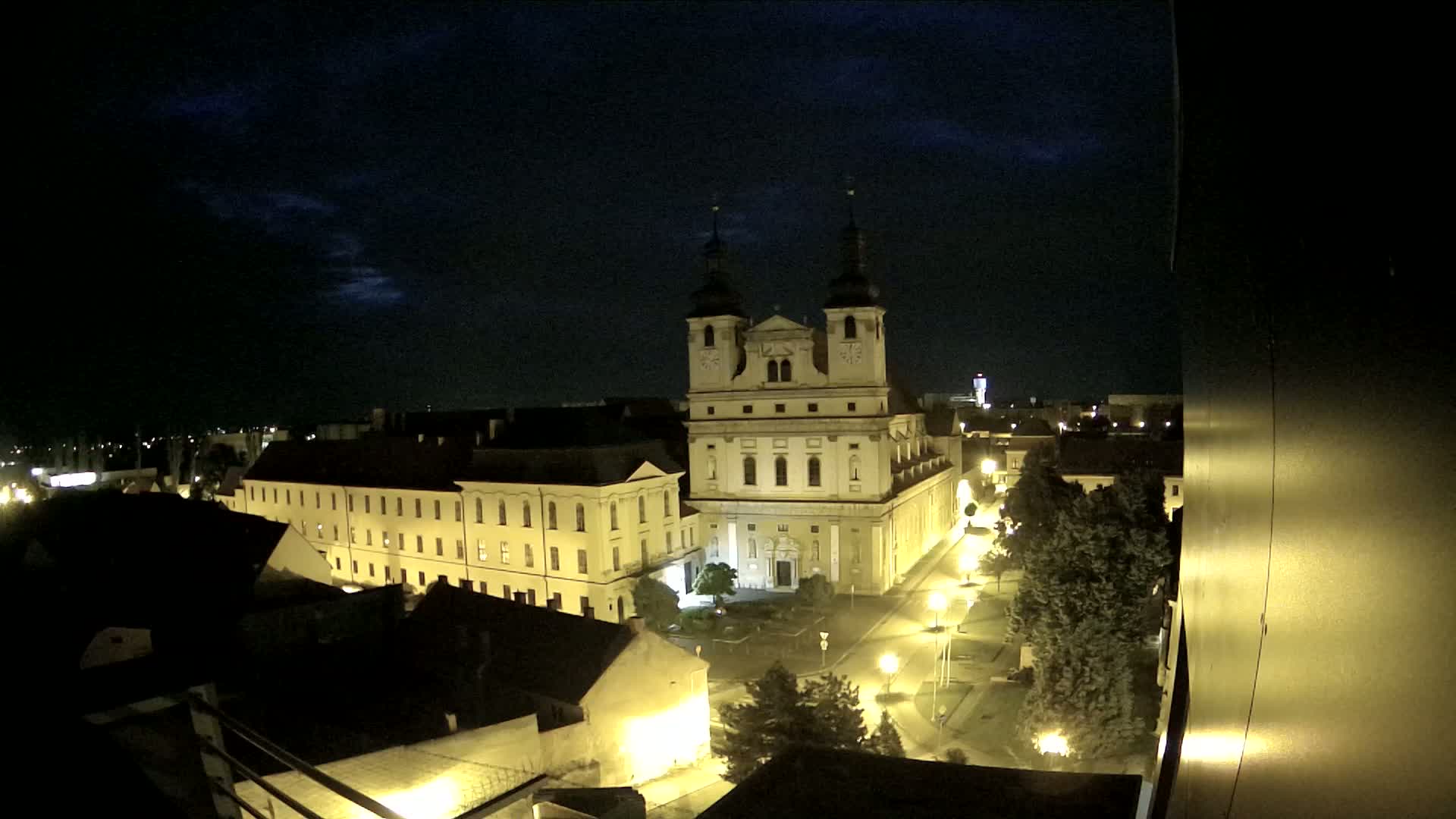 A nighttime view of a city square with a large church illuminated, under a partly cloudy sky.