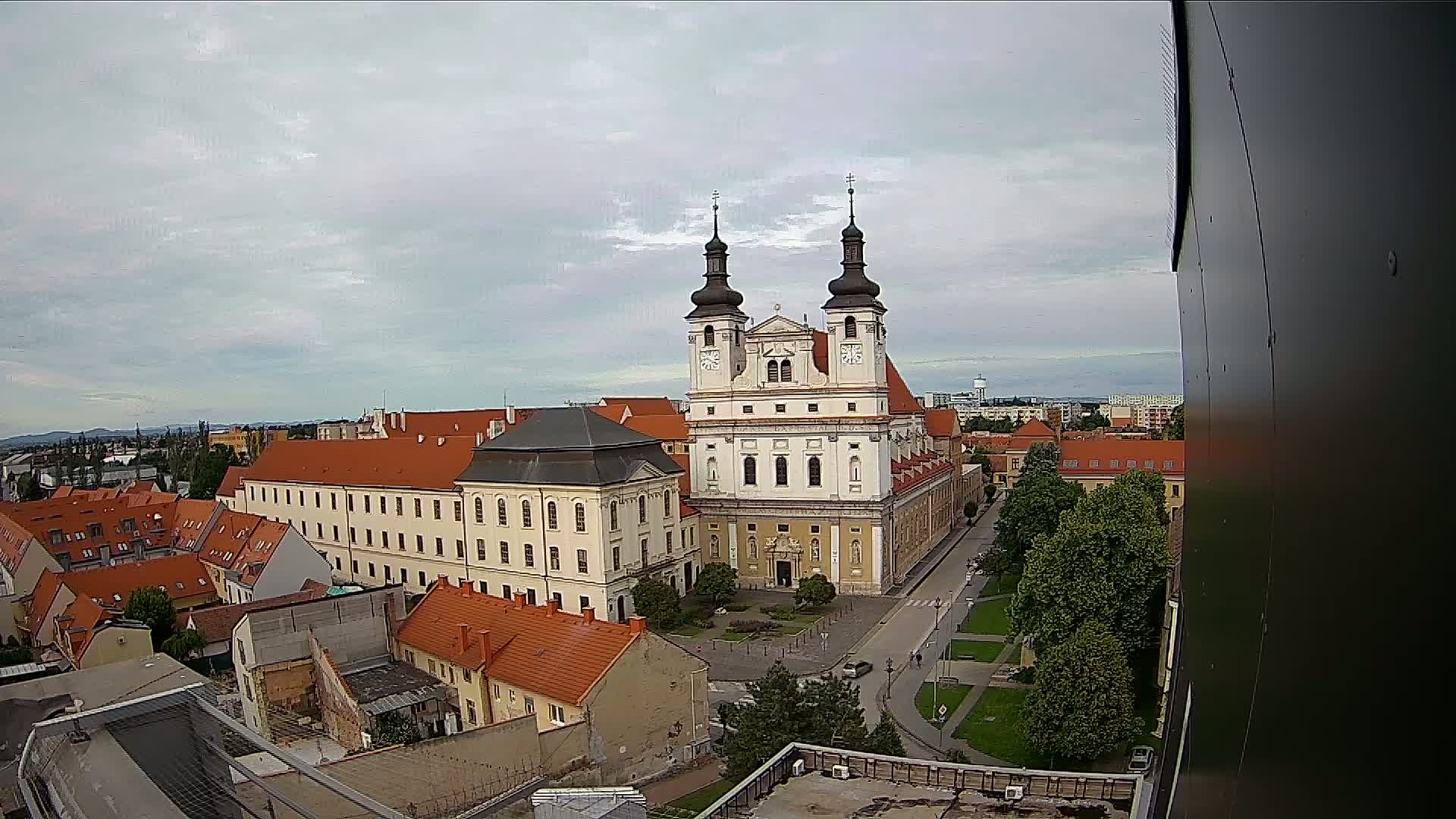 A large, light-colored church with twin towers stands in a city square, surrounded by smaller buildings with red-tiled roofs under a cloudy sky.