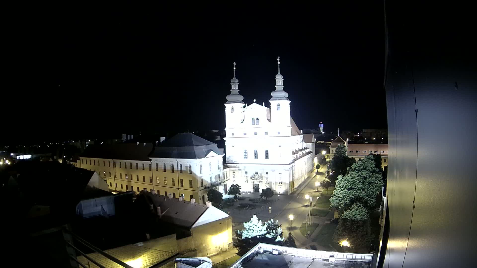 A brightly lit church stands in a town square at night, surrounded by other buildings and trees under a clear, dark sky.