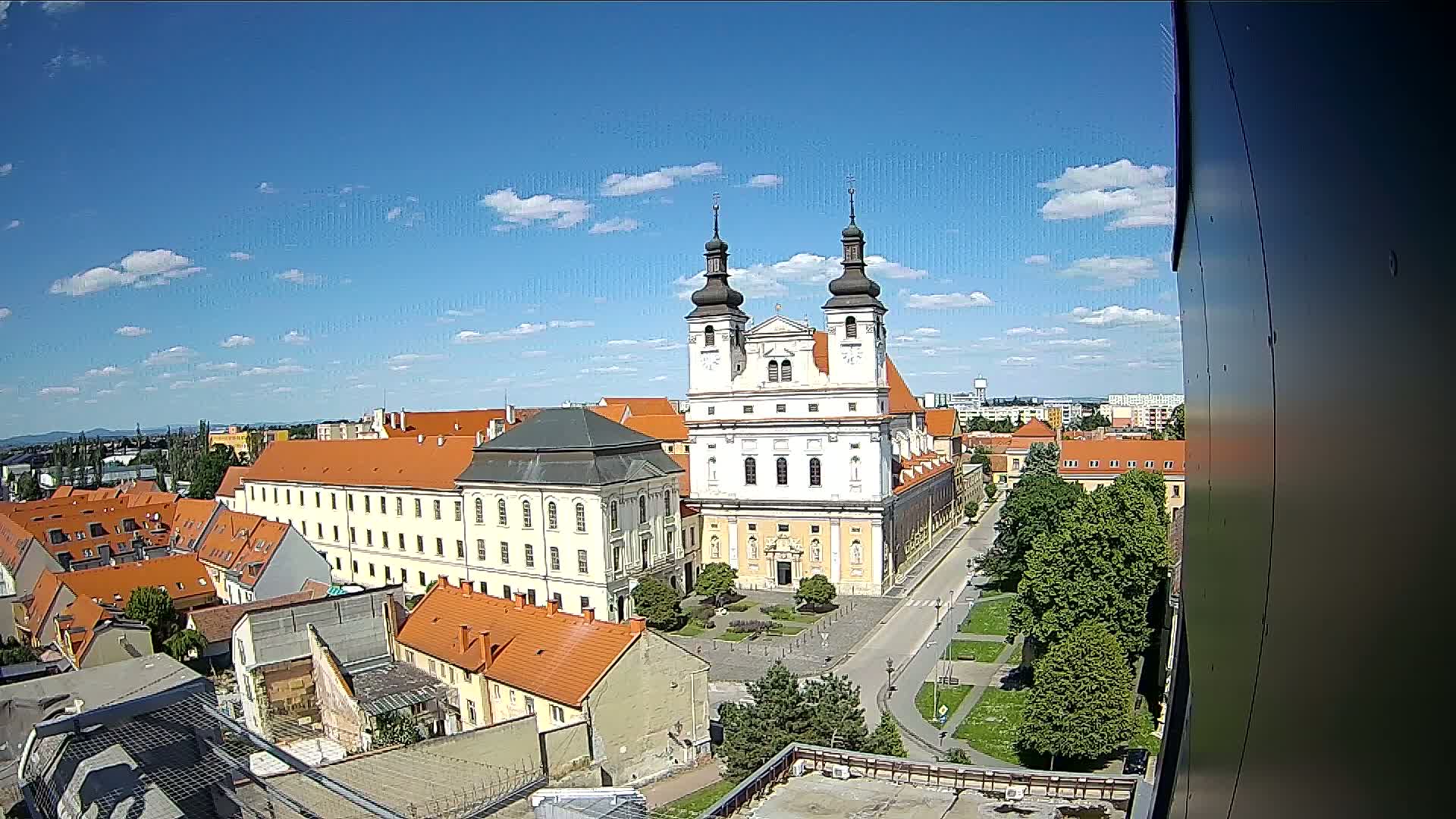 A large, light-colored church with twin towers stands prominently in a city square on a sunny day with partly cloudy skies, surrounded by red-roofed buildings.
