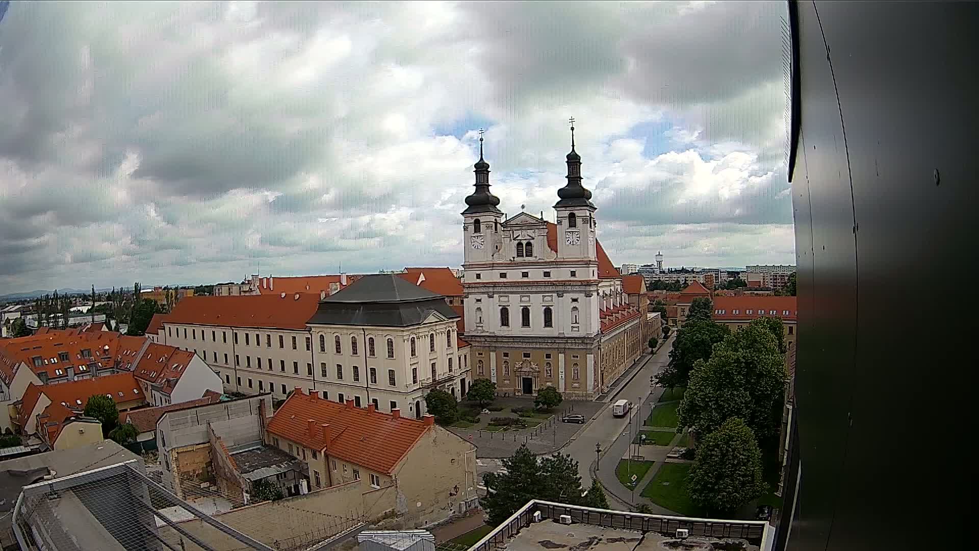 A large church with twin towers is the centerpiece of a city square, viewed from above on a partly cloudy day.