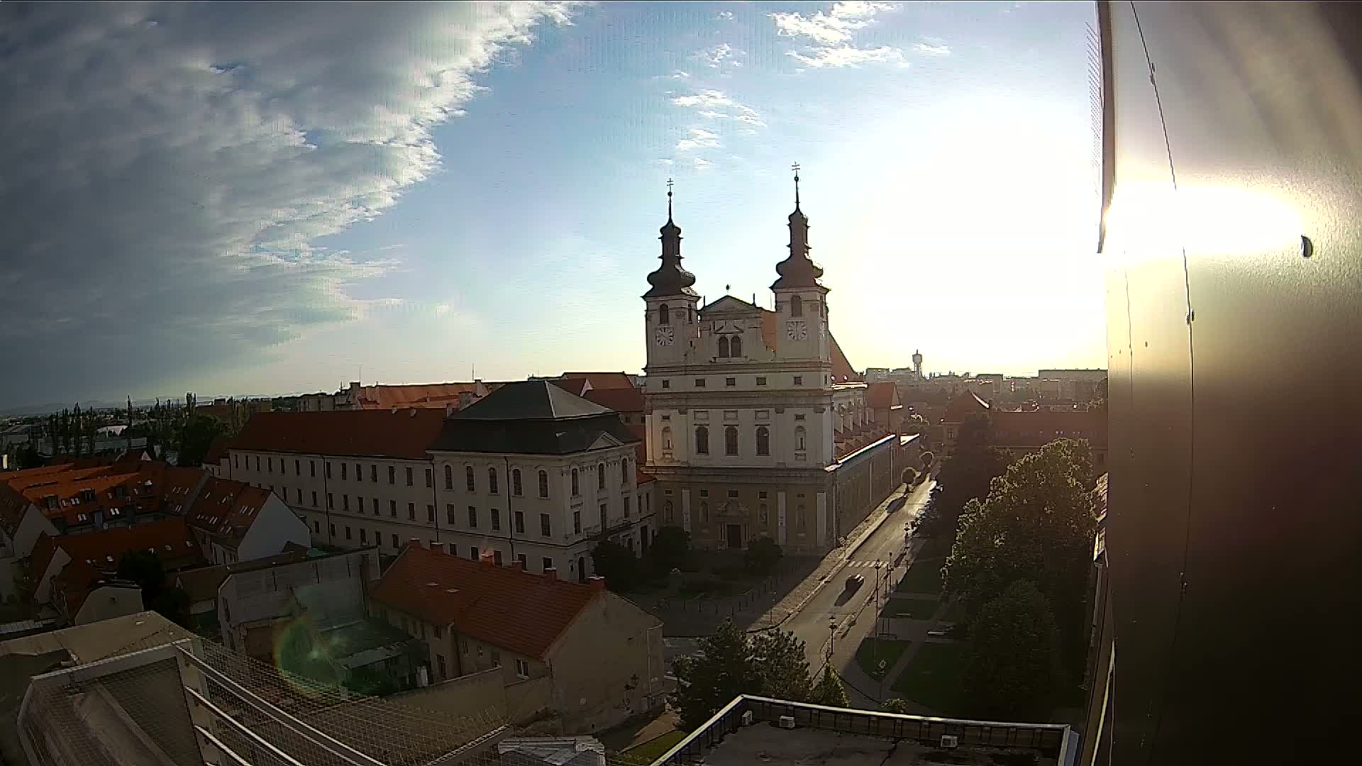 A large church with twin towers stands prominently in a city under a partly cloudy sky, with the sun shining brightly.