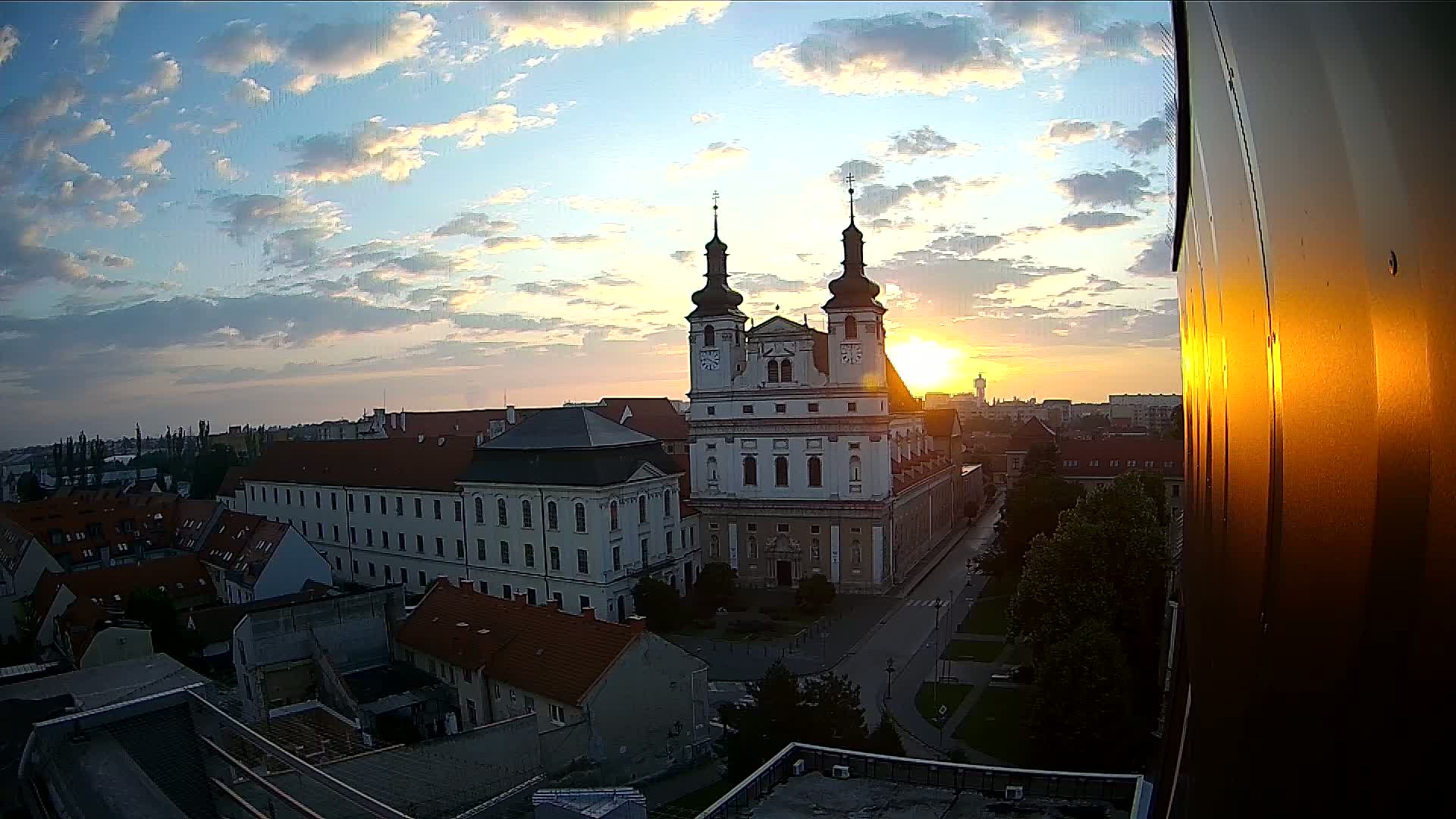 A sunrise illuminates a large church and surrounding cityscape, with a partly cloudy sky.