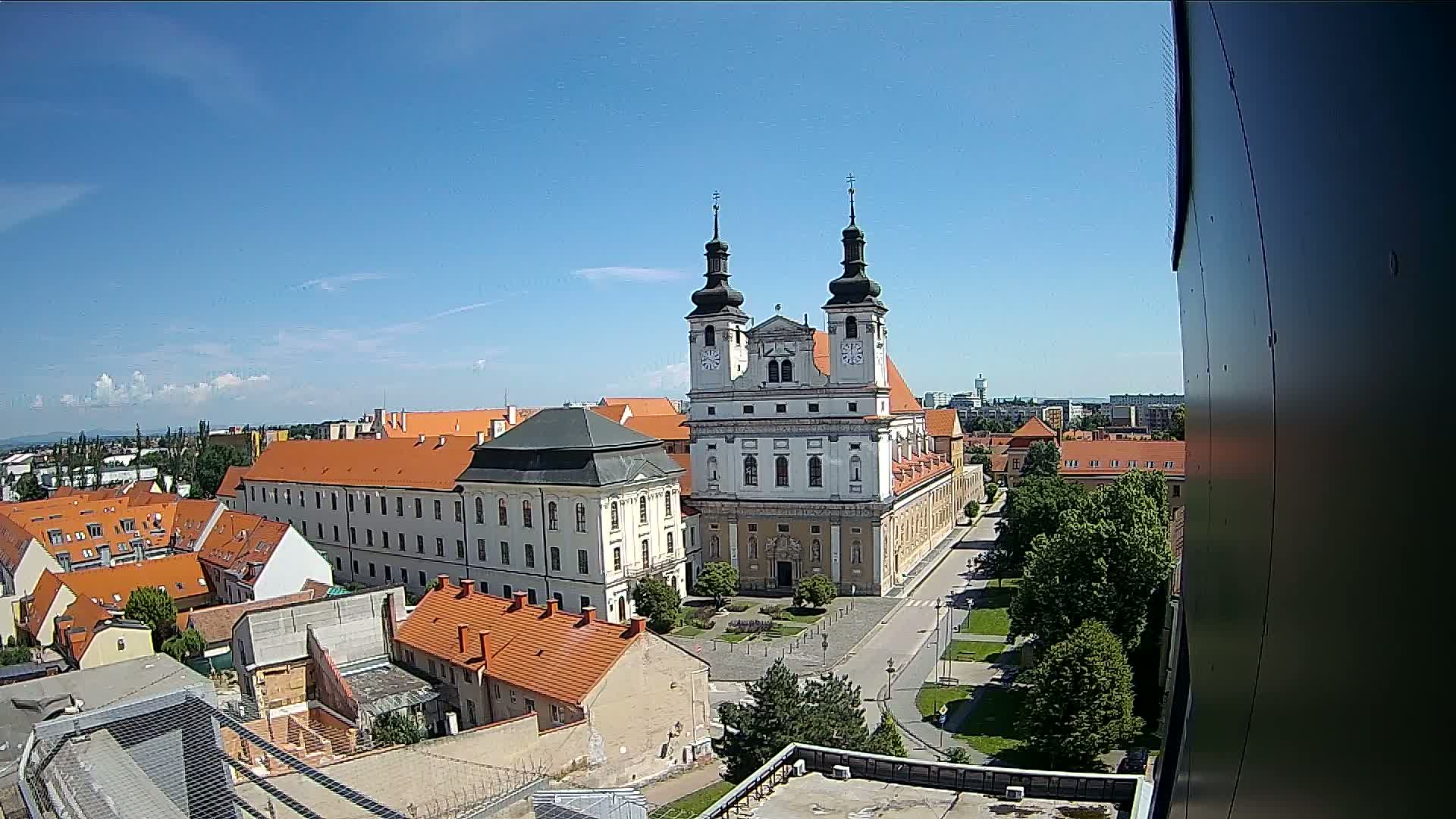 A large, white church with two towers stands prominently in a city square, surrounded by buildings with orange tile roofs under a clear blue sky.