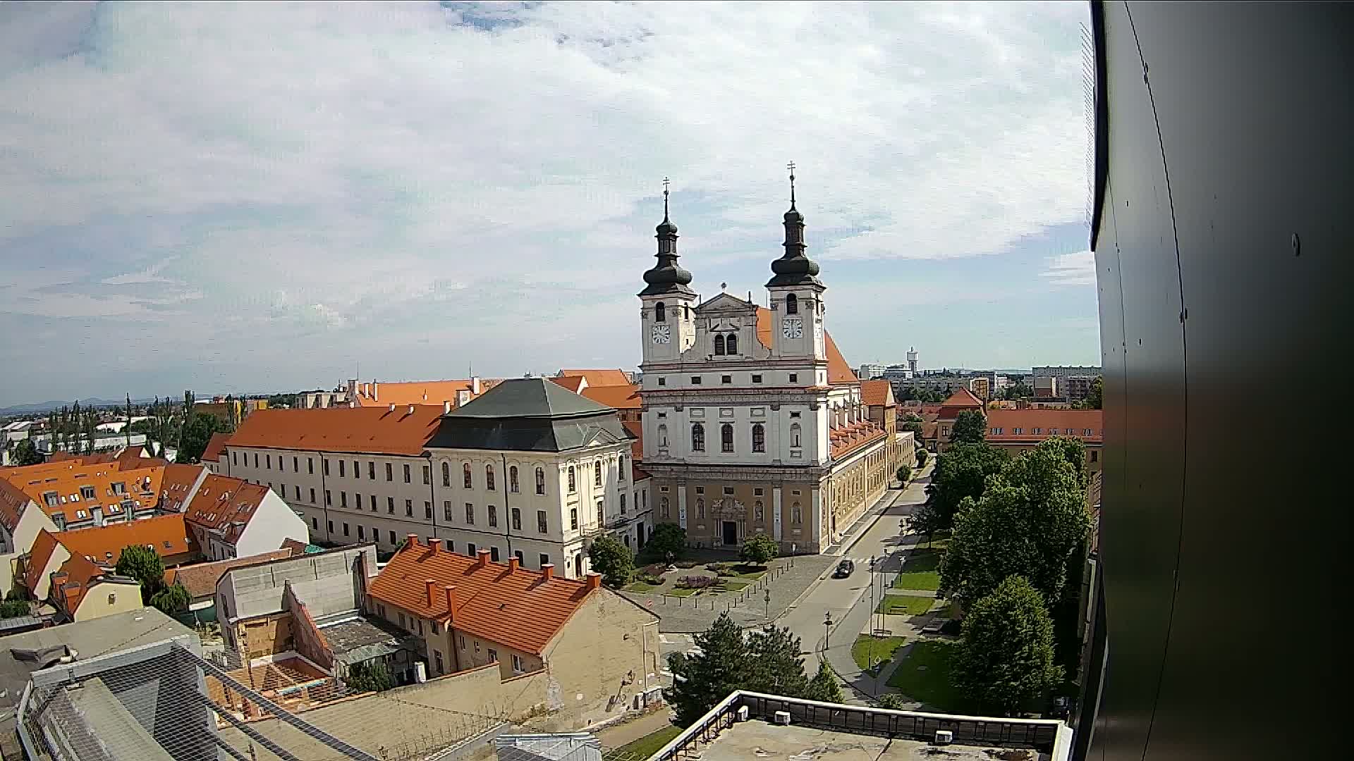 A high-angle view on a partly cloudy day shows a large church with orange-roofed buildings surrounding a town square.