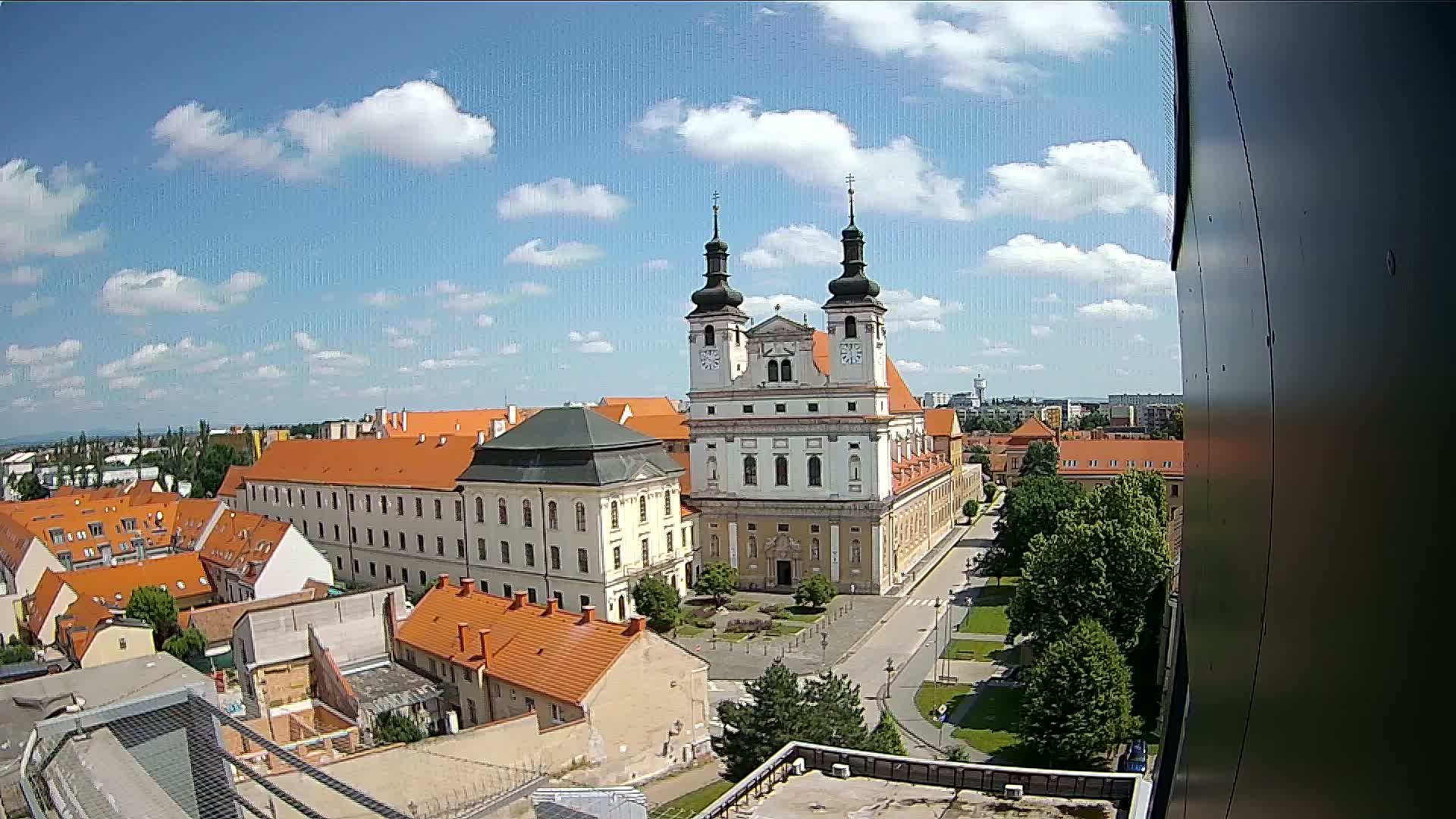 A large church with twin towers and orange-roofed buildings surrounding a town square on a partly cloudy day.