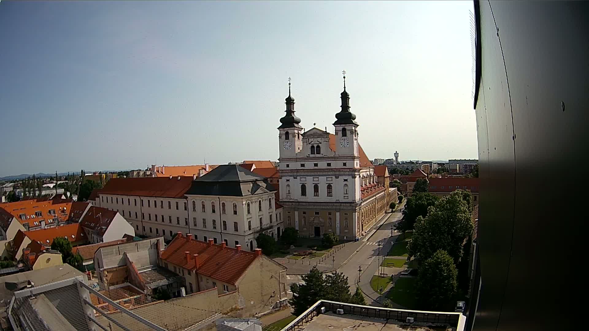A large white church with two towers stands prominently in a city square on a sunny day, surrounded by smaller buildings and green spaces.