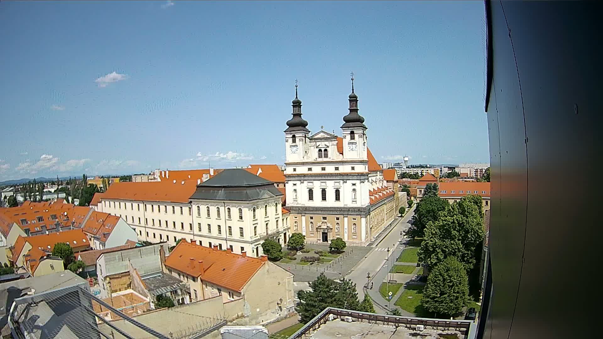 A large church with two towers stands prominently in a city square surrounded by low buildings with orange roofs, under a clear, sunny sky.