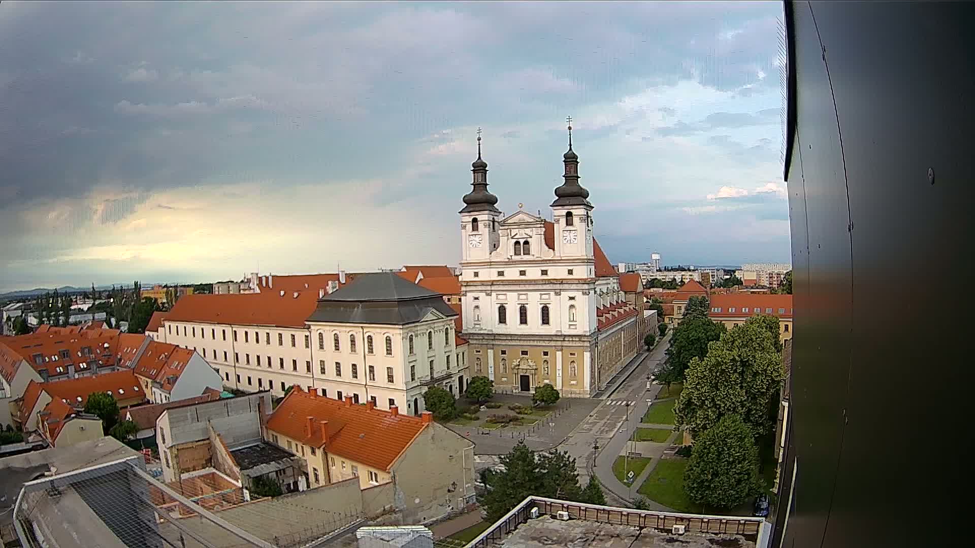 An elevated view of a European city on a partly cloudy day shows a large church complex surrounded by red-tiled buildings.