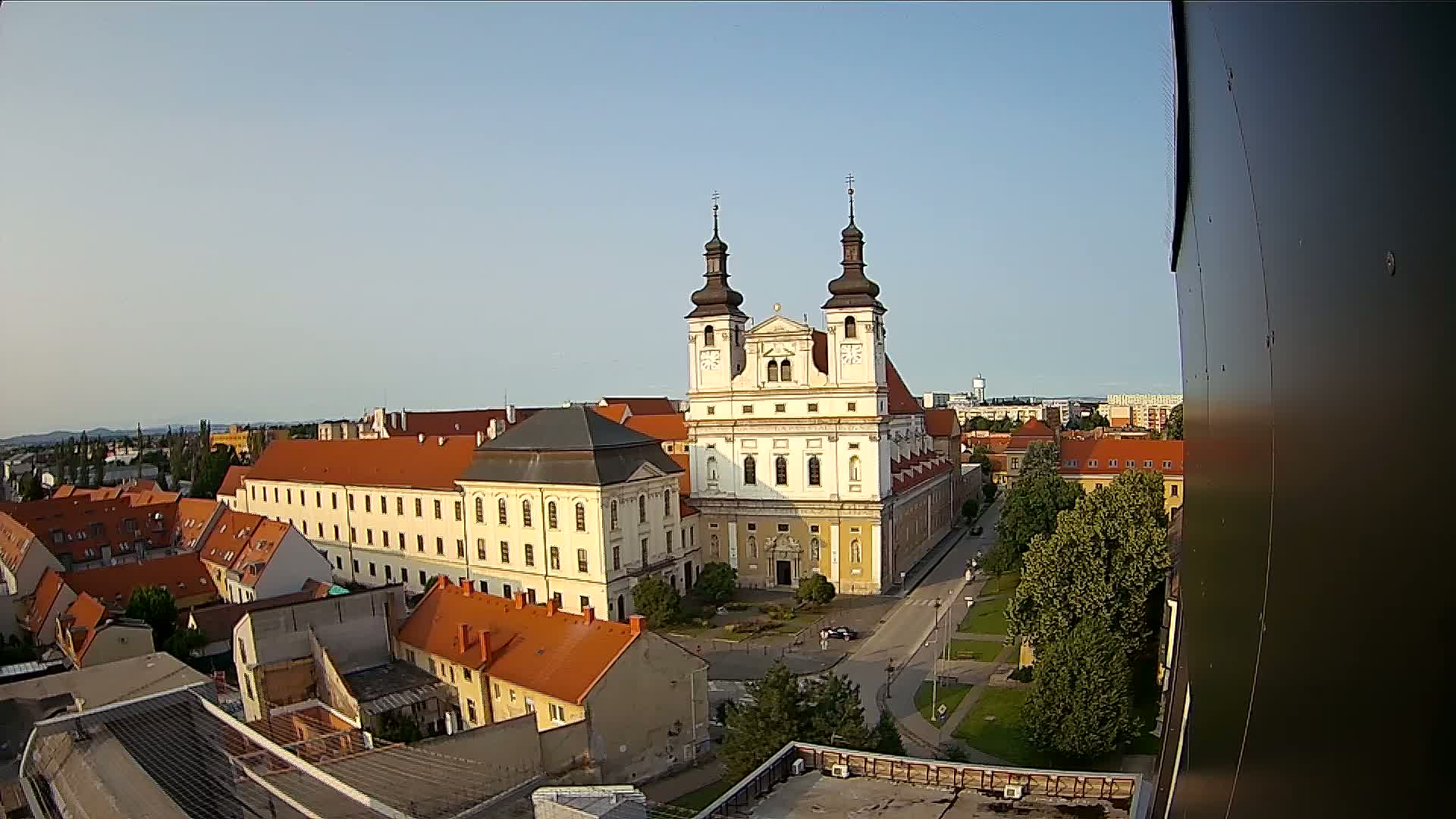 An elevated view of a European town square on a clear, sunny day, centered around a large, light-colored church with twin towers and surrounded by low-rise buildings.