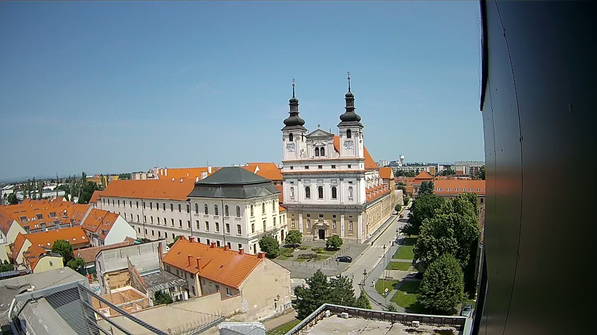 A high-angle, sunny-day view shows a large church with two towers surrounded by buildings with orange-tiled roofs and green trees.