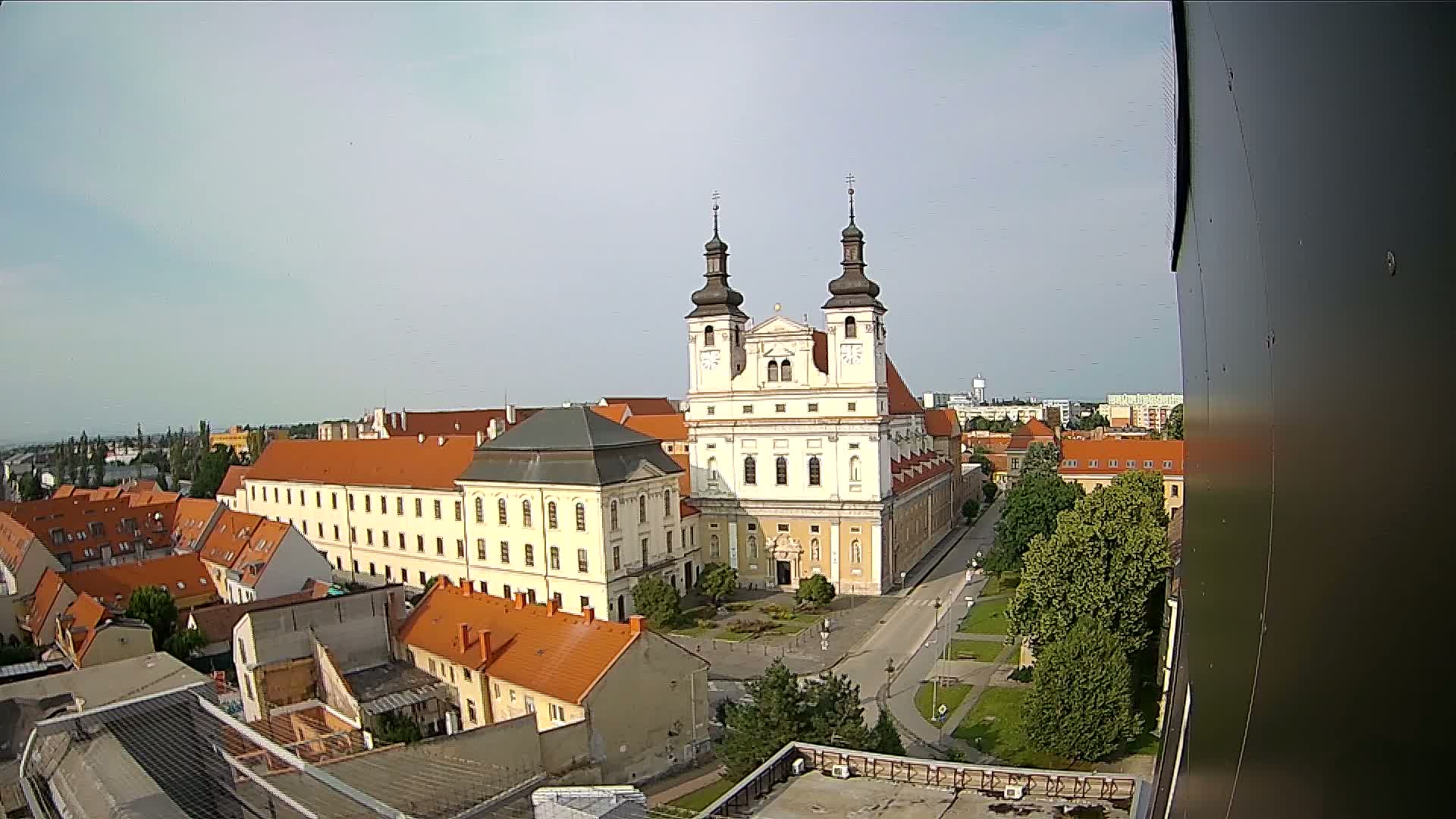 A high-angle view on a partly sunny day shows a large church with two towers surrounded by low buildings, green spaces, and a street.