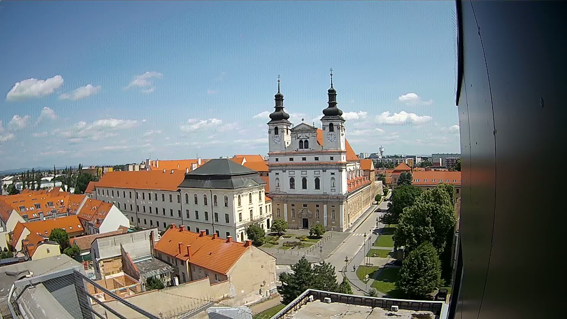 A large church with two towers stands prominently in a city square under a mostly sunny sky with scattered clouds, surrounded by buildings with orange-tiled roofs and green trees.