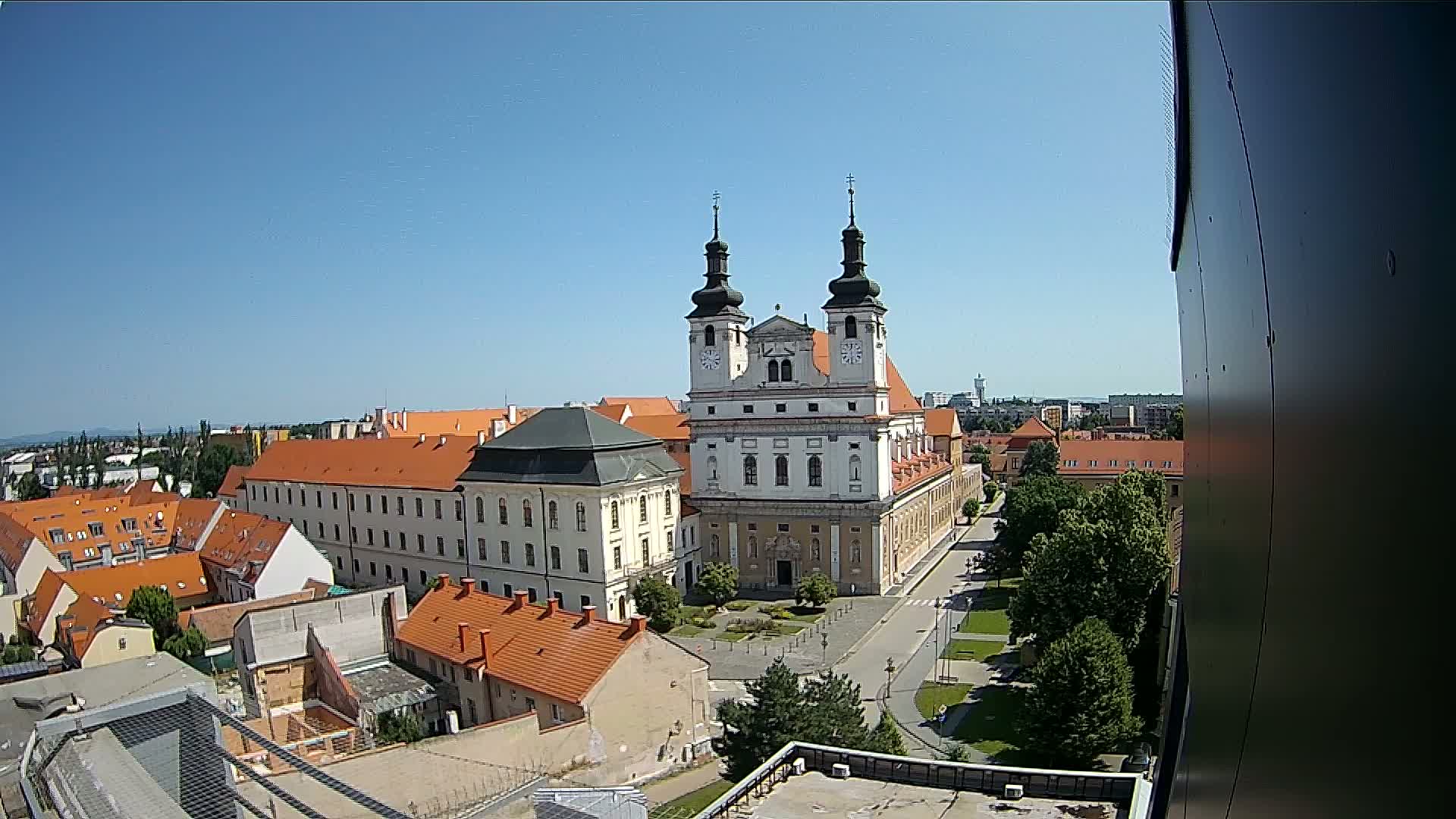 A large church with twin towers stands prominently in the center of a European city on a sunny day, surrounded by low buildings with orange roofs and green trees.