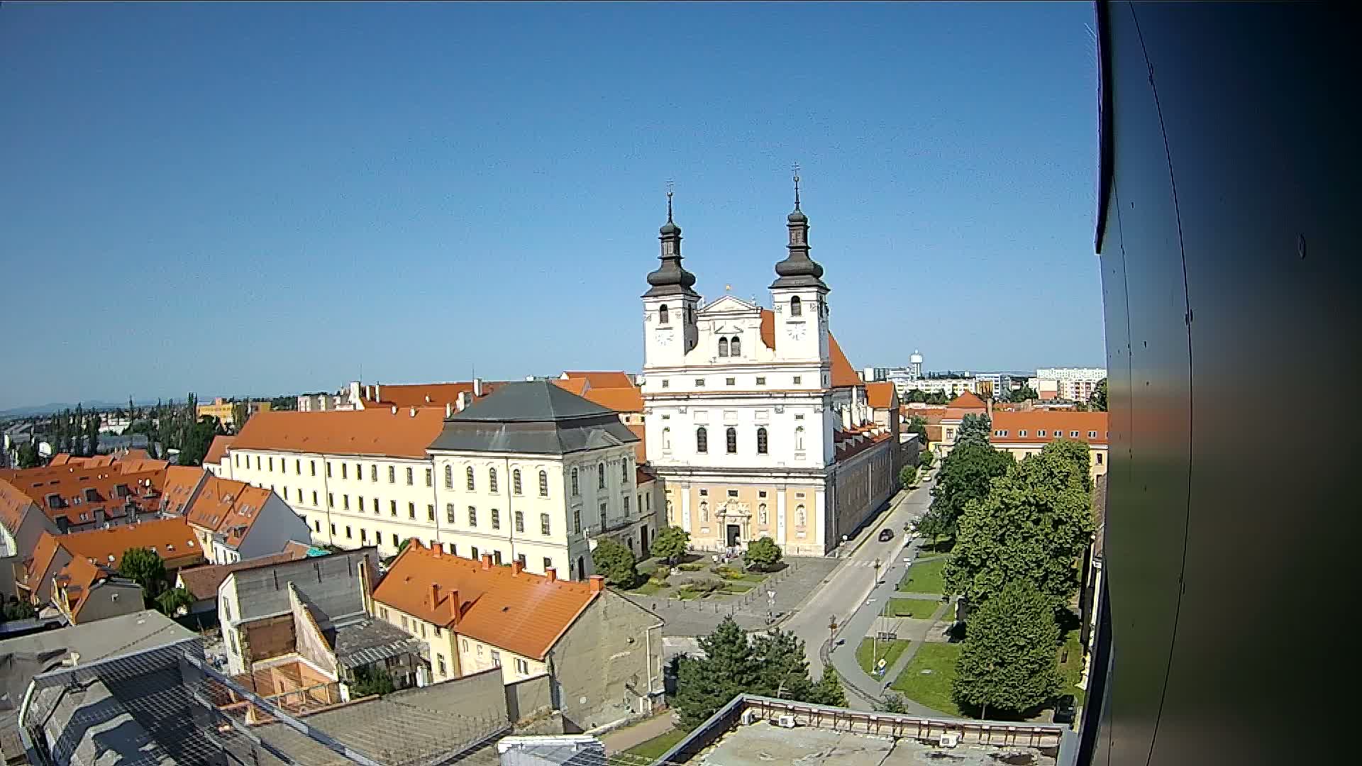 A large, white church with twin towers stands prominently in a town square on a clear, sunny day, surrounded by buildings with orange-tiled roofs.
