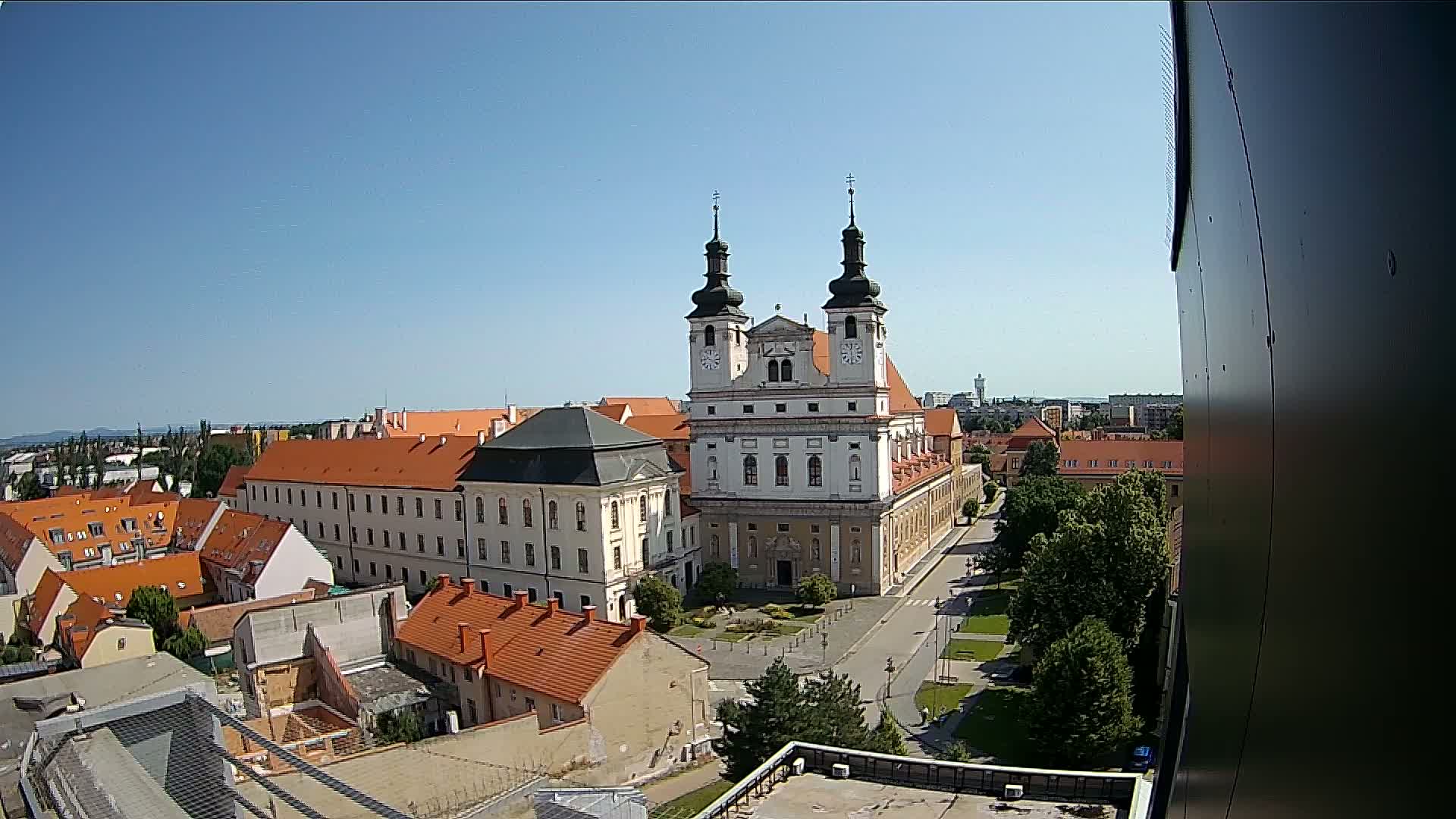A large white church with two towers stands prominently in a town square on a sunny day, surrounded by smaller buildings with orange-tiled roofs and green trees.