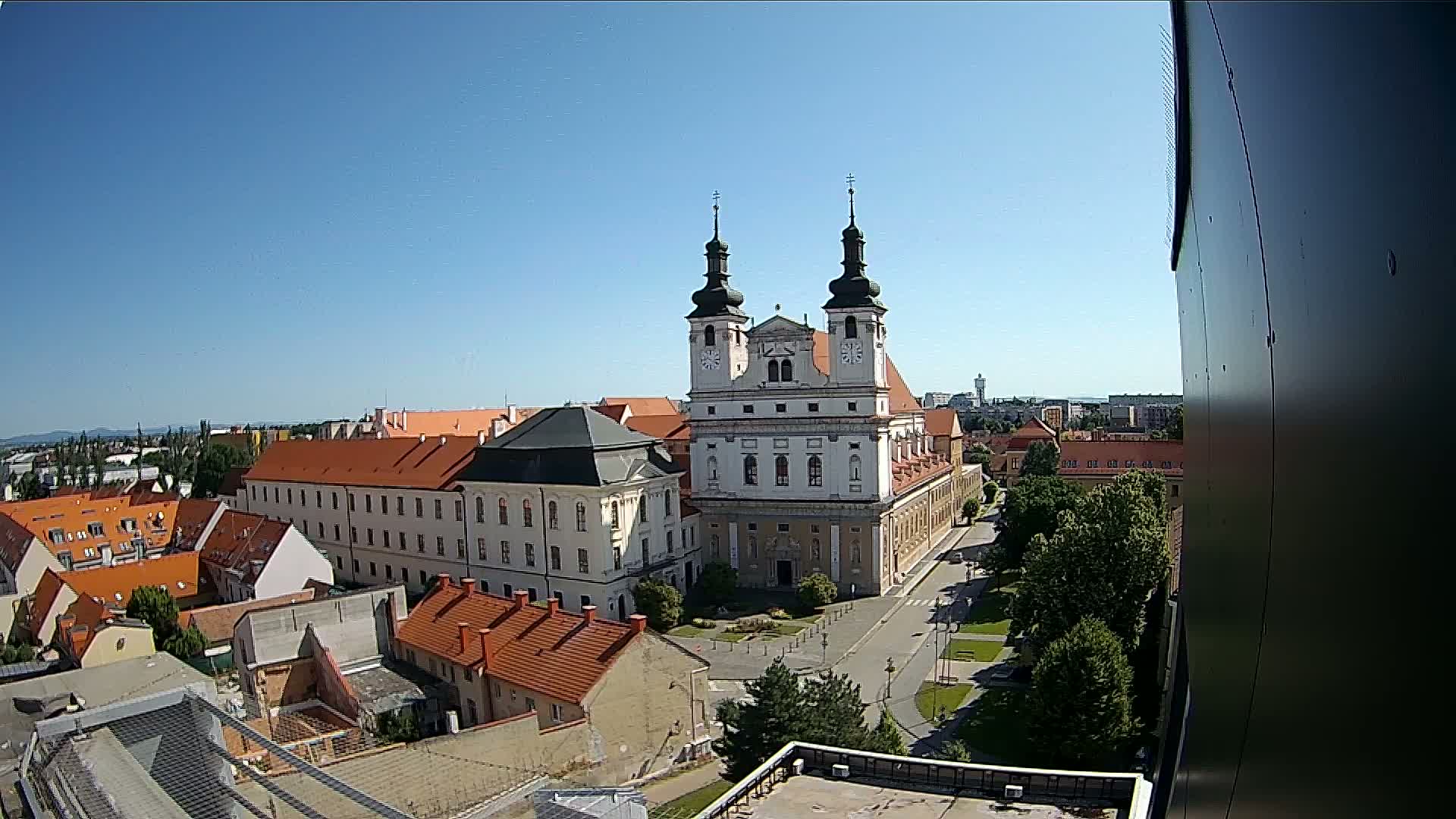 An elevated view on a clear, sunny day shows a large church with twin towers surrounded by a town square and other buildings with red tile roofs.